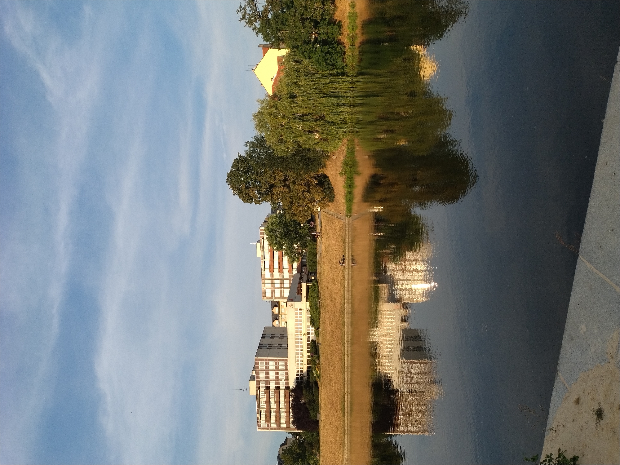 The image shows a daytime scene of the Otava River in Písek, Czechia. On the far bank, several multi-story buildings with white and beige facades are visible, along with green trees. One of the buildings is a light yellow color and has a red roof. The buildings are reflected in the water. The foreground shows the river's surface, which is dark and reflective. The sky is blue with some white clouds. A portion of a concrete embankment is visible in the lower left corner of the image. Two figures are seated on a bench near the buildings on the far bank.