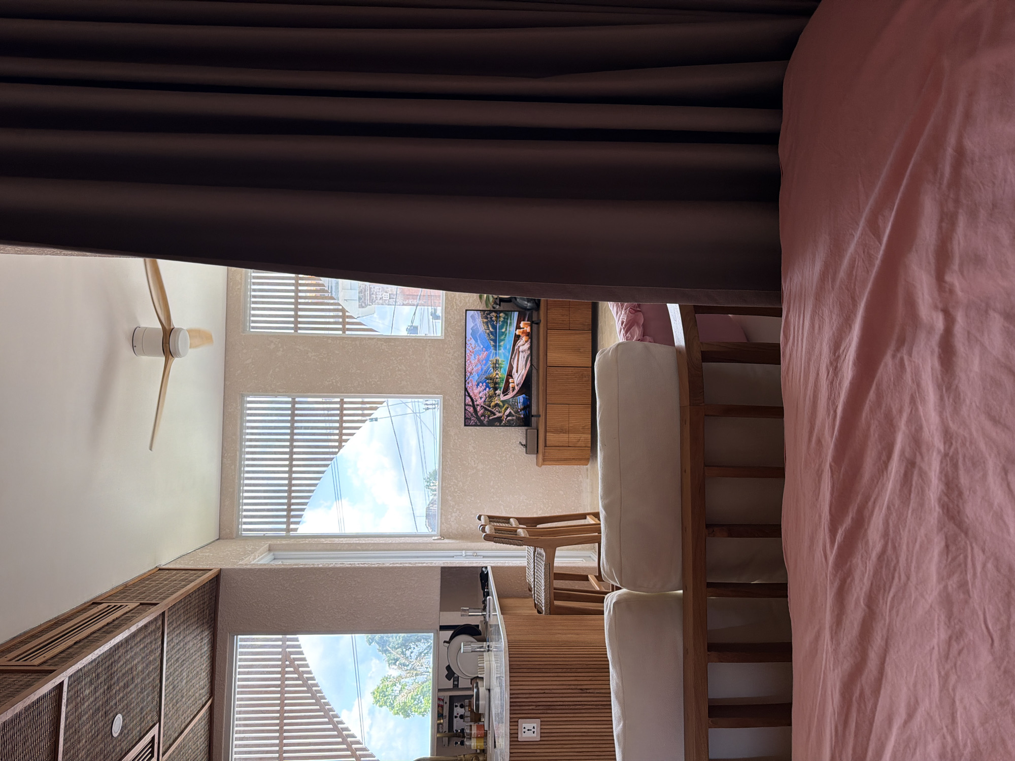 A sideways interior view features a bedroom transitioning into a living area with kitchen elements. Natural light floods in through multiple windows covered with horizontal blinds, revealing exterior views of a bright sky and foliage. The bedroom section includes a large pink fabric and a wooden frame. Adjacent to the bedroom, the living area features a television mounted on a wall above a wooden cabinet. Further back, a kitchen area is visible with a countertop, appliances, and various kitchen items. The image includes a ceiling fan and brown curtains along the top. The location is possibly in Mengwi, Indonesia, based on the building style, architecture, and foliage visible through the windows.