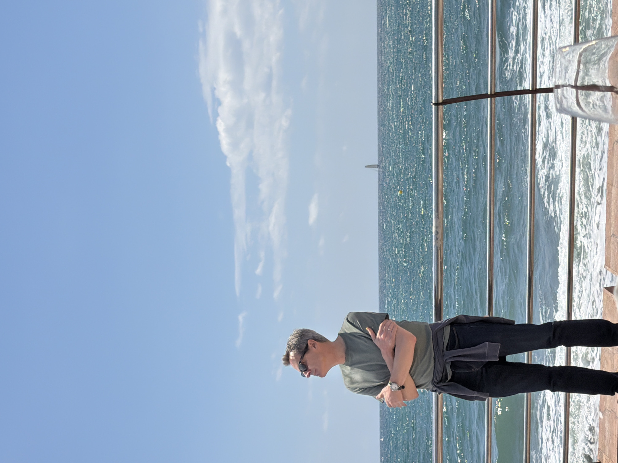 A medium-shot captures a man standing by a seaside railing in Arroyo de la Miel-Benalmádena Costa, Spain. The man, wearing sunglasses, a green shirt, black pants, and a jacket tied around his waist, has his arms crossed. The railing separates him from the sea, which is sparkling under the sunlight. The sky above is blue with scattered clouds. The scene has a calm, touristy feel.