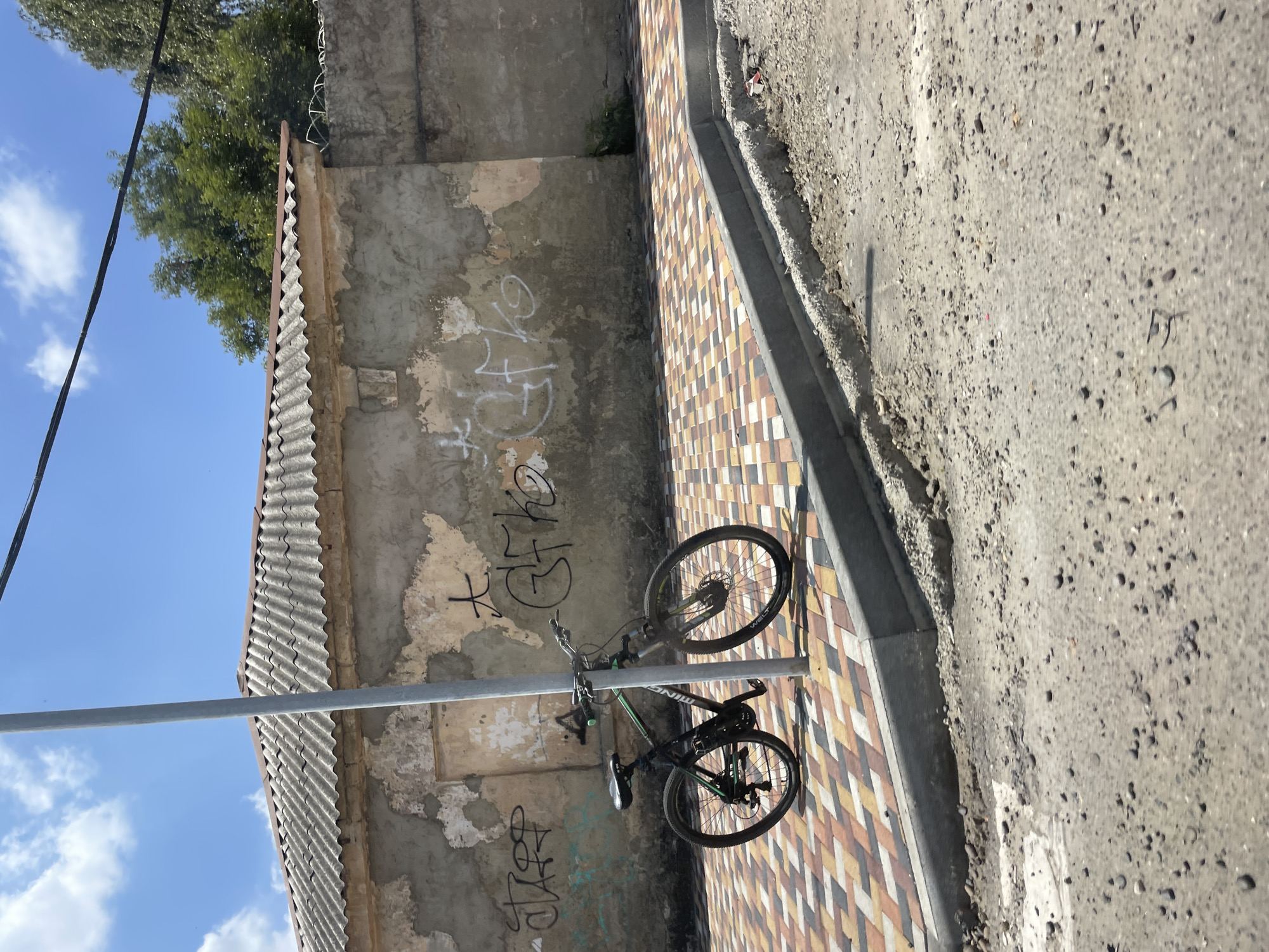 An outdoor, eye-level, medium shot captures a bicycle leaning against a metal pole in an urban setting. The bicycle, black with green accents, is positioned on a colorful, patterned sidewalk. Behind the sidewalk is a weathered, beige wall covered in graffiti, topped by a corrugated metal roof. The sky above is blue with scattered white clouds. The foreground shows a rough, gravel-covered area, suggesting ongoing construction or repair work. The scene appears to be in the city of Stavropol, Russia. There are no people visible in the image.