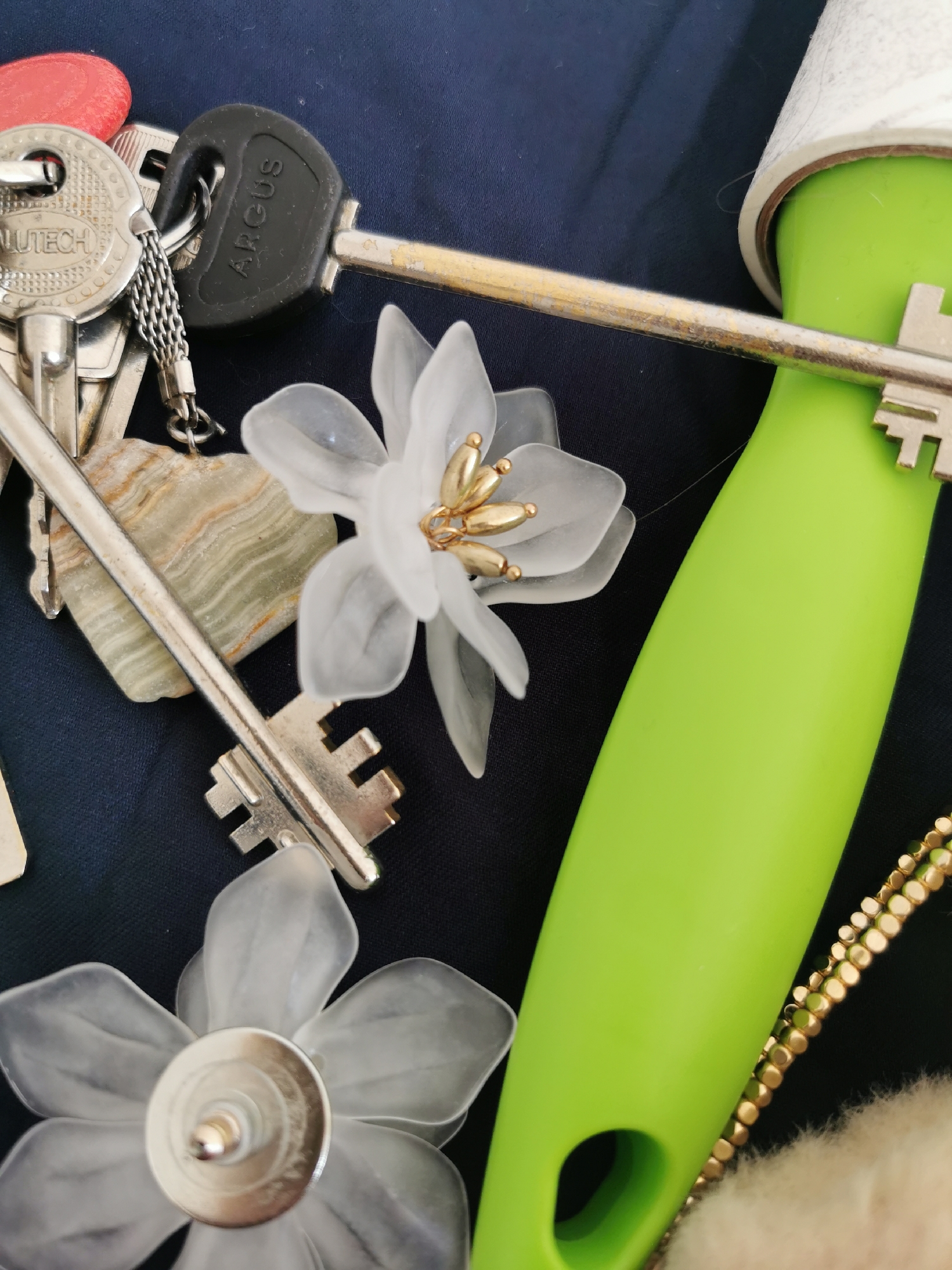 The image shows a collection of objects on a dark blue surface. There are several keys, including one with a red tag, one with the brand "Lutech", and one with the brand "Argus". There is also a light green lint roller, two white flower-shaped objects that appear to be earrings, a stone pendant, and a gold beaded chain. The scene does not provide enough information to determine a specific location in Moscow, Russia.