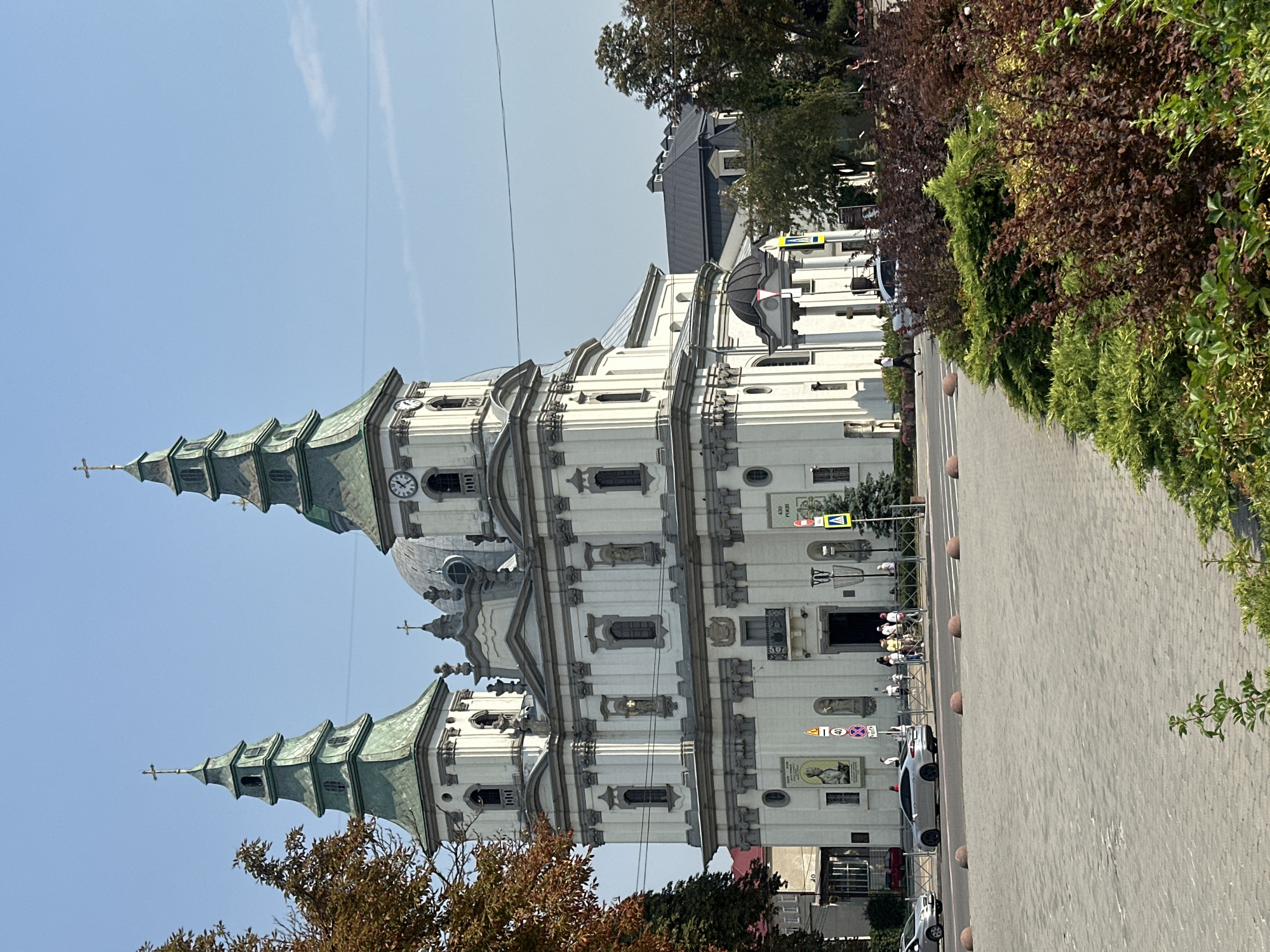 A wide shot captures the Ternopil Cathedral of the Immaculate Conception in Ternopil, Ukraine, bathed in bright sunlight under a clear blue sky. The grand, white baroque church features two prominent green-roofed towers topped with crosses, and a large dome. A clock is visible on the left tower. The facade is adorned with decorative architectural elements, statues, and windows.
In the foreground, a paved plaza leads to the church entrance. Several people are gathered on the sidewalk in front of the cathedral, some appearing to be tourists or parishioners. Cars are visible on the street, including a silver sedan and a white SUV. A pedestrian crossing signal and traffic signs are also present.
The scene is framed by lush green trees and shrubs lining the plaza and sidewalk, with some autumnal foliage visible on the left. The overall impression is a vibrant urban setting with a significant religious landmark.