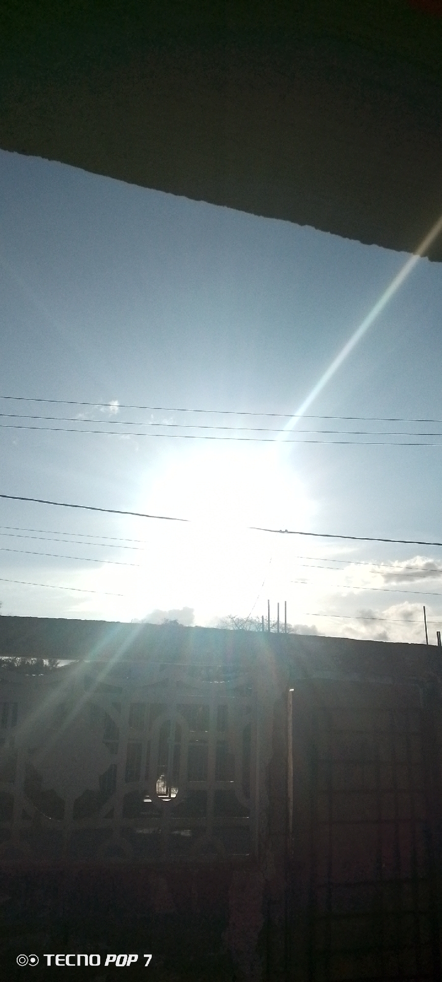The image shows a bright, sunny day in Altagracia, Venezuela. An unidentifiable dark object obscures the top portion of the frame. Below, a light blue sky is visible, with the sun shining brightly and creating a strong glare. Several power lines run horizontally across the sky. Below the power lines is a dark, silhouetted rooftop, with some vegetation and structures (possibly chimneys) visible behind it. In the foreground, there's a dark fence with ornate, patterned openings and possibly a building or wall. The image is somewhat underexposed. The text "TECNO POP 7" is visible in the bottom left corner.