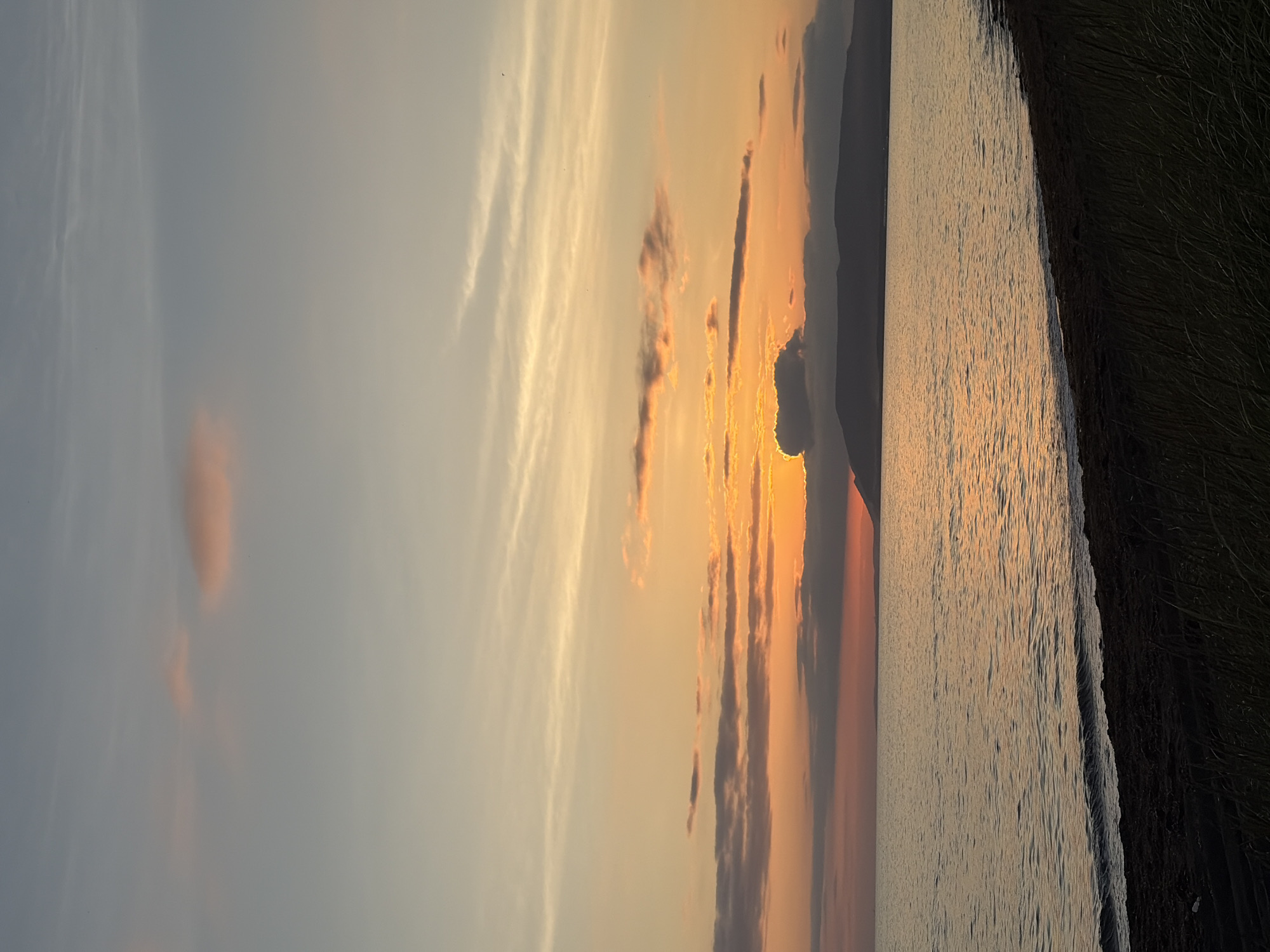A serene sunset over the sea at Ozerskoe, Russia, paints the sky with hues of orange, pink, and pale blue. Wispy clouds drift across the horizon, some tinged with the sun's warm glow. The calm sea reflects the vibrant sky, with gentle waves lapping at a dark, sandy shore. Silhouetted against the fading light, a distant mountain range forms a dark, undulating line across the seascape. Tall, dark grasses line the foreground, adding a touch of texture to the natural landscape. No people or animals are visible in the image.