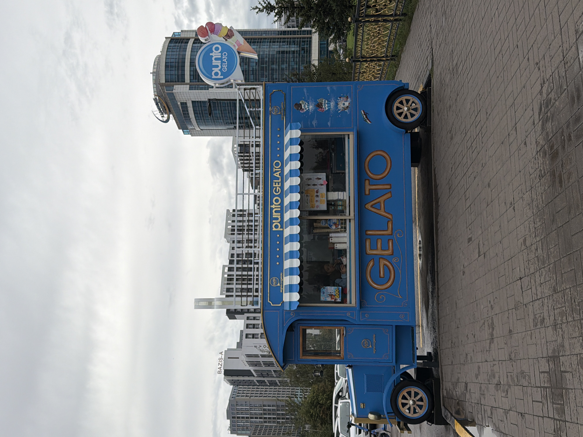 An outdoor, eye-level, full shot captures a vibrant blue gelato truck parked on a wet, brick-patterned plaza in Astana, Kazakhstan. The truck, designed to resemble an old-fashioned vehicle with ornate gold lettering spelling out "GELATO," features a serving window and an awning. Inside the window, a person is visible, appearing to be working. Above the truck, a circular blue sign with white text reads "punto GELATO" accompanied by an illustration of ice cream cones. In the background, modern high-rise buildings dominate the skyline, with one particularly tall, glass-fronted structure bearing a circular logo and text at its peak. A decorative gold metal fence runs along the right side of the plaza. The sky is overcast and appears cloudy. Cars are visible parked behind the gelato truck to the left.