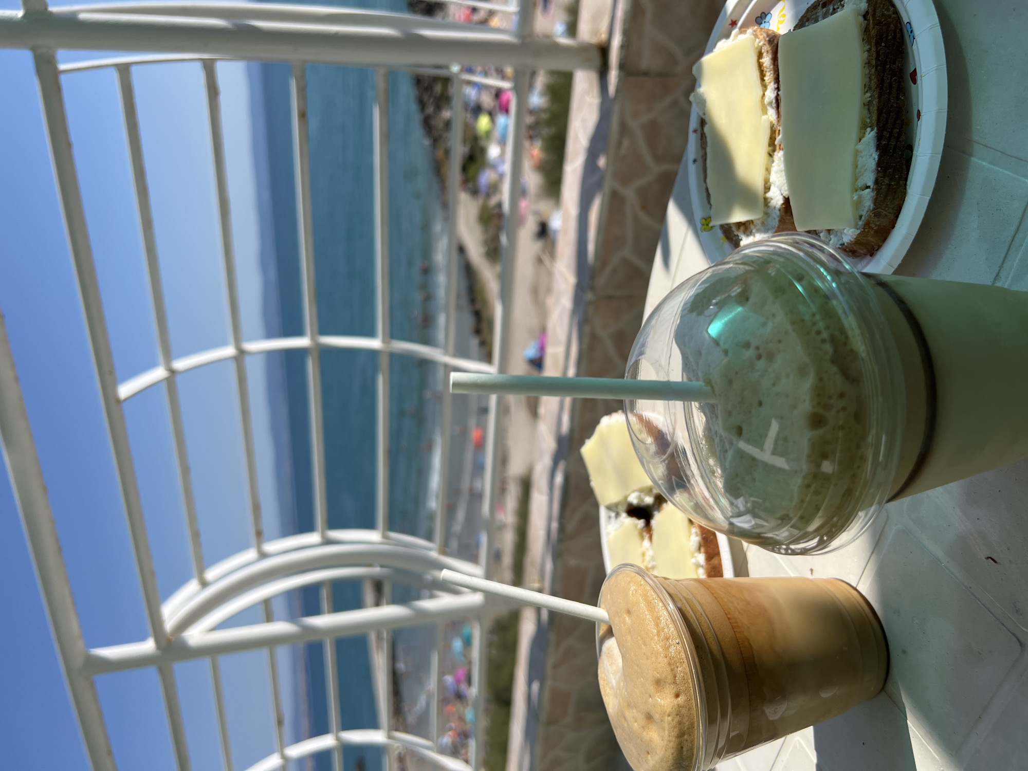 The image depicts a breakfast scene on a table with a white railing in the foreground and the sea in the background. Two glasses of iced coffee are placed on the table, one in a clear plastic cup and the other in a transparent plastic cup with a lid. Next to the coffee cups, a plate holds two slices of bread topped with a white spread and cheese. In the background, the sea is visible, with a beach dotted with umbrellas and people. The overall scene suggests a relaxed morning by the sea in Pomorie, Bulgaria.