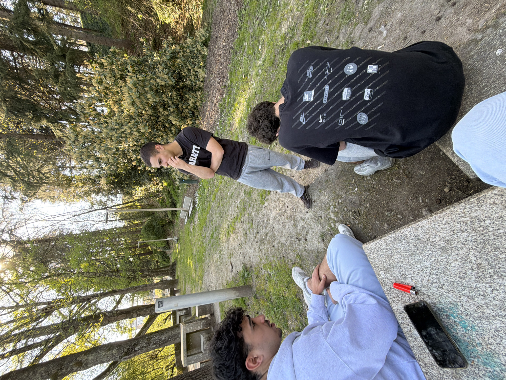 The image shows a group of young men outdoors in Vila Real, Portugal. Three people are visible in varying degrees of detail. One man is sitting on a concrete bench, wearing a light-colored hooded sweatshirt and light-colored pants. A red lighter and a black smartphone rest on the bench next to him. Another man is standing on a dirt/grass area, wearing a black t-shirt with white writing, and grey pants. A third man is partially visible, wearing a black t-shirt with Nike logos on the back. The setting appears to be a park with trees and benches. The ground is covered in dirt, grass, and scattered debris.