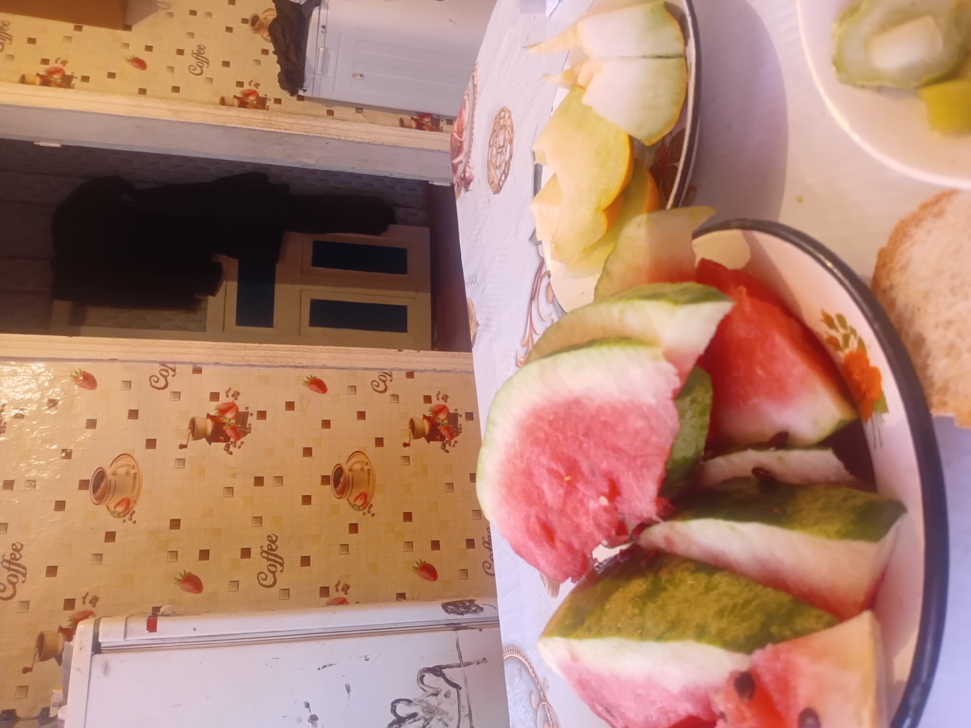 The image depicts an indoor scene, likely a kitchen or dining area, with a table set with various fruits. Slices of watermelon and melon are arranged on plates, suggesting a meal or snack is being served. The background features wallpaper with a repeating pattern of coffee cups, coffee grinders, and strawberries. To the left, a white refrigerator is visible, and a doorway in the background reveals a dark interior with a hanging garment. The overall impression is one of a casual, domestic setting. The provided location, Малая Владимировка ауылдық округі, Kazakhstan, provides a geographical context for this scene.