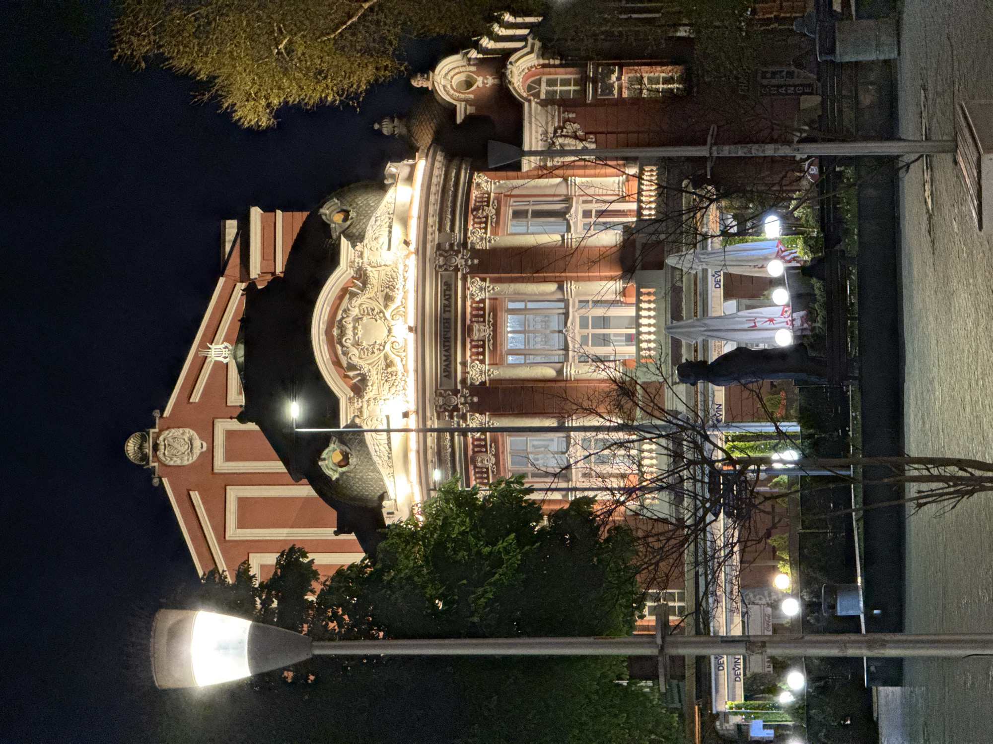 The image shows the Drama Theater "Stoyan Bachvarov" in Varna, Bulgaria, at night. The building is ornately decorated and illuminated. A statue is partially visible to the right, along with outdoor café umbrellas and street lamps. Trees are present in the foreground, partially obscuring the lower portion of the building. The ground is paved with light-colored stones.