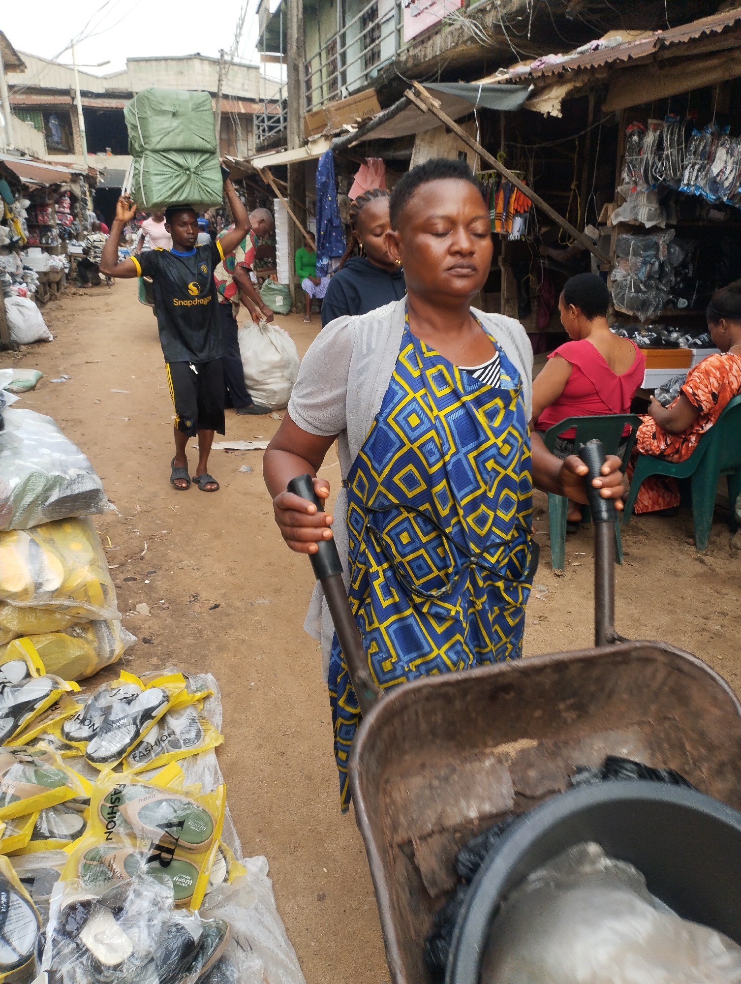 The image depicts a bustling scene in what appears to be the Fegge area of Onitsha, Nigeria, characterized by informal markets and commercial activity.
A woman is the primary subject, pulling a rusty wheelbarrow filled with items contained in a black plastic bag and a white plastic bag. She is wearing a blue and yellow apron-like garment over a striped shirt, covered in a light gray sweater.
In the background, a man carries a load of green bags on his head while walking along a dirt path lined with stalls and shops. People are seated at stalls, possibly engaged in selling or other business activities. The structures in the background appear to be a mix of corrugated metal and basic construction, suggesting a market or commercial area. There are also plastic bags filled with sandals visible on the ground to the left.