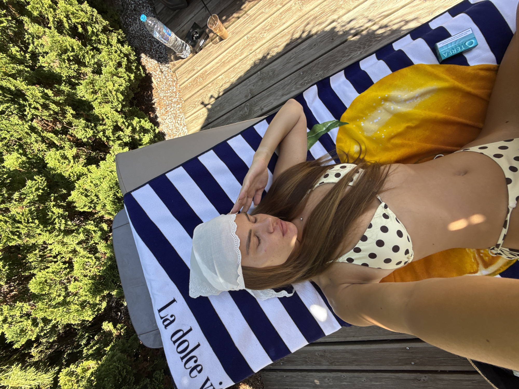 A woman is lying on a lounge chair on a wooden deck, outdoors. She is wearing a polka dot bikini and a white bandana. The lounge chair has a striped towel with the words "La dolce vita" on it and a large lemon print. A bottle of water, a glass with ice, and a small package are on the deck next to the chair. In the background, there is lush green foliage. The scene appears to be in a private backyard or garden area.