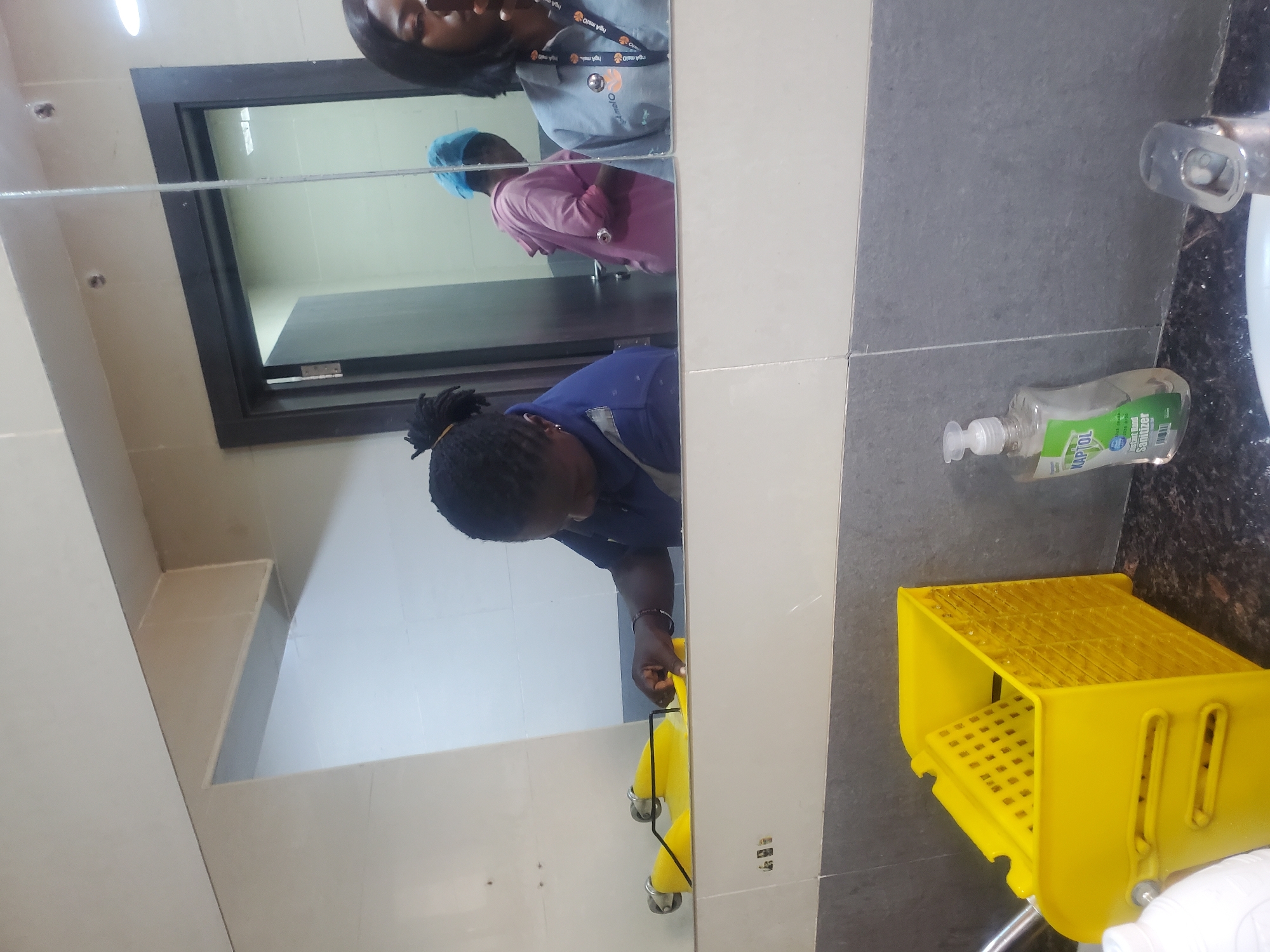The image, rotated 90 degrees clockwise, captures an indoor scene within a bathroom or restroom in Gwagwada, Nigeria. The central feature is a large mirror reflecting several individuals and parts of the room.
In the reflection dominating the foreground, a person with dark, tied-back hair and wearing a blue shirt is actively cleaning. Their right hand is visible, holding a yellow cloth or sponge, wiping a surface, likely the mirror or the counter beneath it. Adjacent to this activity, on the right side of the image, is a bright yellow cleaning bucket equipped with a wringer.
Further back in the mirror's reflection, two other people are visible. One individual, positioned towards the top left of the reflection, is wearing a light grey top and a lanyard with a discernible "Oranges" or similar logo. Behind them, a third person is seen in a pink top, wearing a light blue cap or hairnet, standing near a dark-framed doorway.
On the tiled countertop to the right of the mirror, a clear plastic bottle of "KAPTOL Hand Sanitizer" is clearly visible, next to a chrome faucet. The walls are composed of light beige/white and grey tiles. The scene suggests a period of facility maintenance or cleaning, possibly in a public or commercial building. The lighting is artificial, characteristic of an indoor setting, with no indications of the time of day.