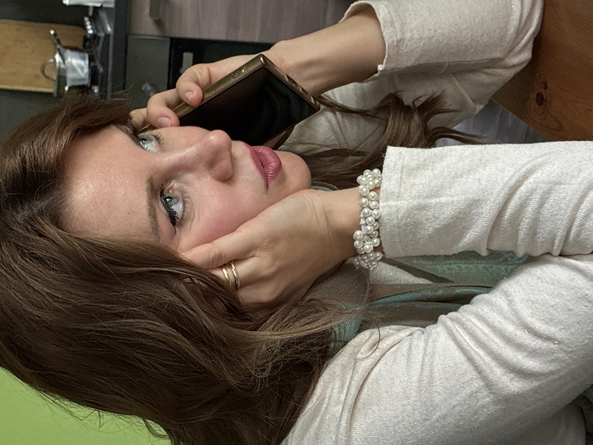 A woman with light skin, brown hair, and blue eyes is holding a smartphone to her ear, seemingly engaged in a phone call. She is wearing a light-colored, long-sleeved shirt and a pearl bracelet. She has gold rings on her fingers. The background is out of focus but appears to be a kitchen area, possibly in a private residence. There is a stainless steel pot visible on the counter in the background, along with some dark cabinetry. There is no explicit visual evidence in the photo to confirm the location to be Moscow, Russia.