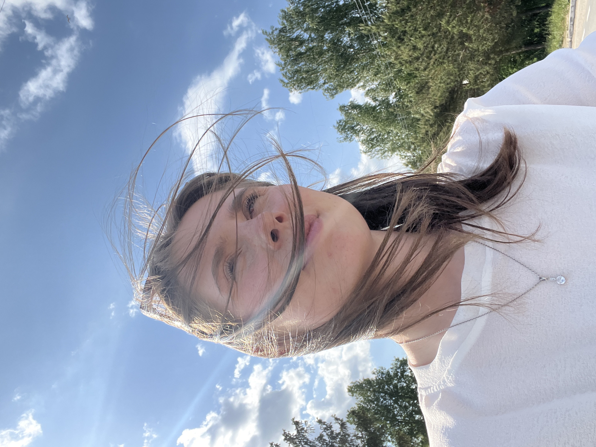 A young woman with long brown hair stands in front of a backdrop of a partly cloudy blue sky and lush green trees. Her hair is disheveled, with strands blowing across her face. She wears a white blouse and a delicate silver necklace. The photograph appears to have been taken from a low angle, pointing upwards. There is no indication of this photo being in Chaikovsky, Russia.