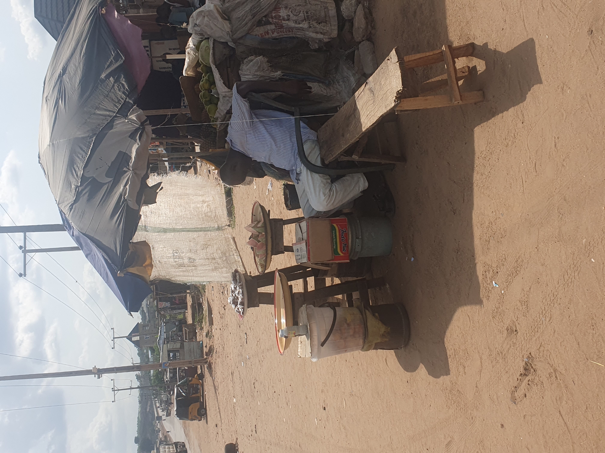 The image depicts an outdoor scene in Lokoja, Nigeria. A man sits on a makeshift wooden chair behind a small stall selling various goods. The stall is shaded by a large, patched umbrella. The items for sale include fruits, packaged food items (a box of "Indomie" instant noodles is visible), and other miscellaneous goods displayed in buckets and on raised surfaces. The man is wearing a striped shirt and light-colored pants. He appears to be attending to his stall. The ground is sandy. The scene is on a street or roadside; in the background, there are buildings, utility poles, and vehicles. The overall setting suggests a street-side market or informal commercial area.
