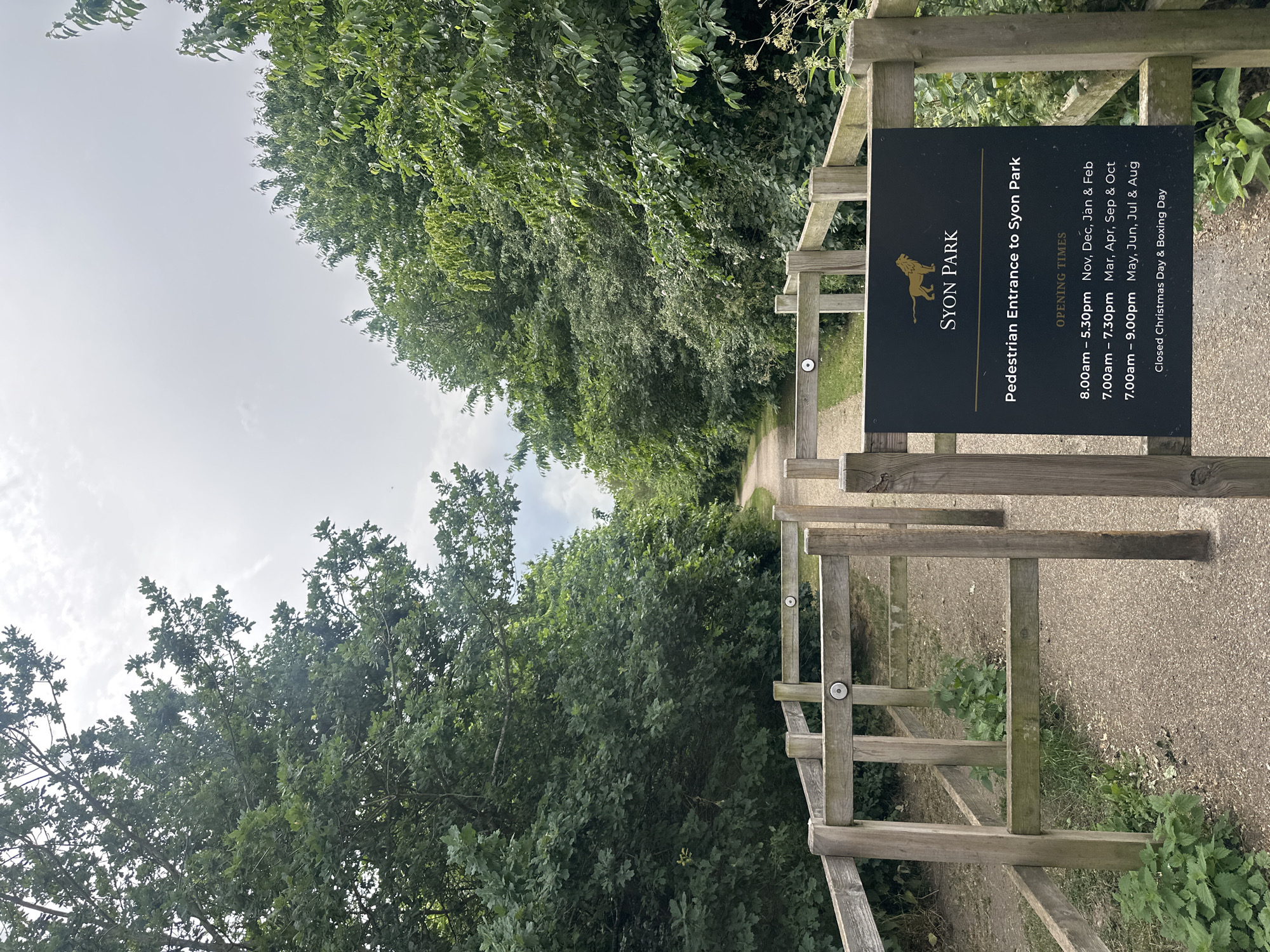 The image shows the pedestrian entrance to Syon Park in Greater London, United Kingdom. A sign displays the name of the park and the opening times throughout the year. A wooden fence surrounds the entrance, leading to a pathway that extends into a wooded area. The sky is overcast.