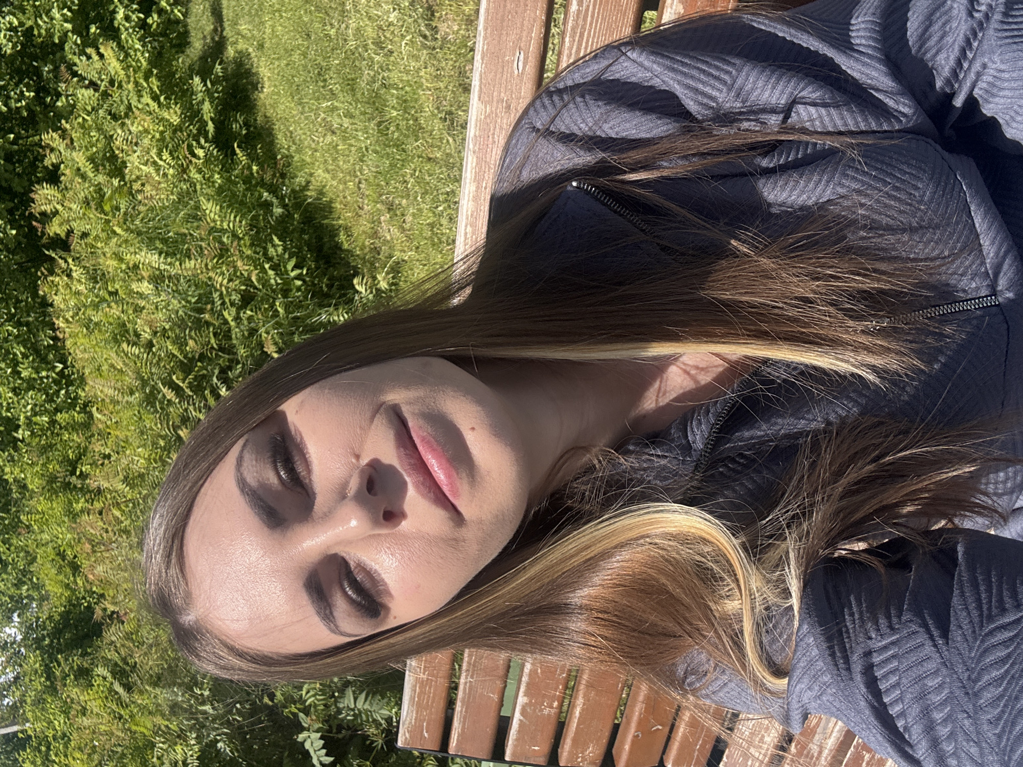 A woman is lying on a wooden bench, possibly outdoors, with grass and foliage visible in the background. The woman has light skin, dark eye makeup, and brown hair with blonde highlights. She is wearing a dark gray zippered jacket with a textured pattern. The scene appears to be lit by natural sunlight. The location cannot be determined as Shevlyakovo, Russia, based solely on the image.