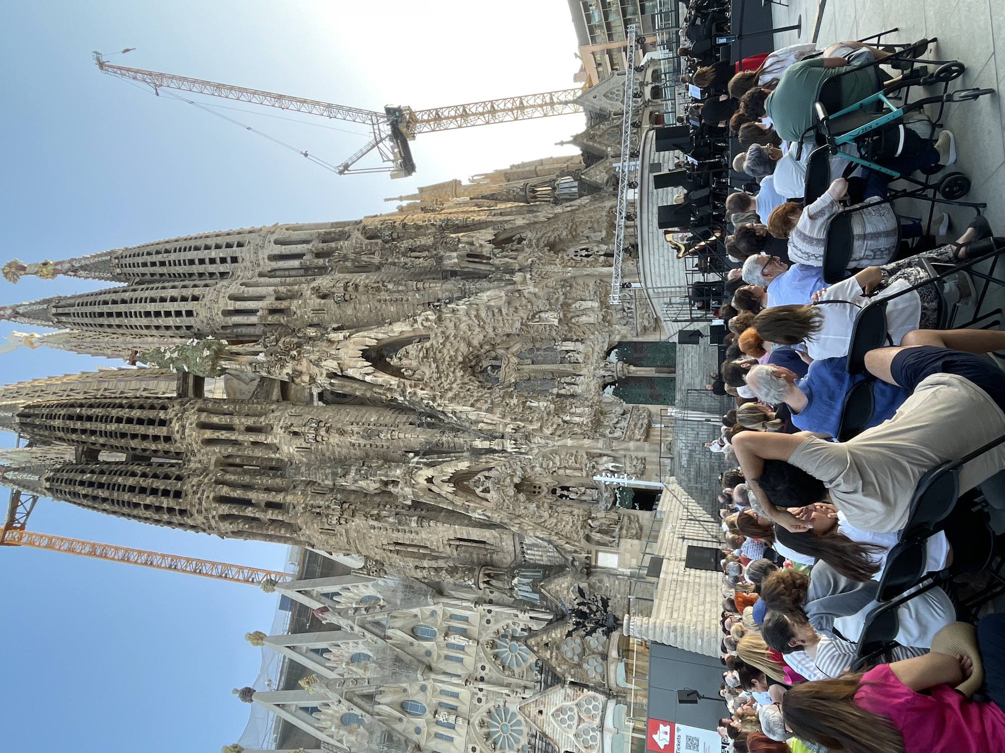 This is a wide-angle, low-angle, outdoor photograph taken in the late afternoon or early evening, depicting a large, ornate, Gothic-style basilica, heavily adorned with intricate sculptures and spires, which is under construction, evidenced by the presence of at least two tall orange construction cranes positioned against a clear blue sky. In the foreground and middle ground, a crowd of people are seated on black folding chairs, facing away from the camera and towards a raised stage area. On the stage, an orchestra is performing, with musicians visible playing various instruments, including what appears to be a harp. The audience appears to be diverse in age and attire. The basilica's facade is incredibly detailed, with numerous figures and architectural elements. The overall scene suggests an outdoor concert or cultural event taking place in front of this iconic landmark. The location is the Sagrada Familia in Barcelona, Spain.