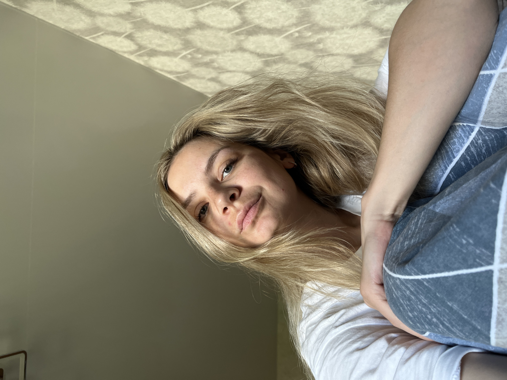 A medium-angle, indoor selfie shows a blonde-haired woman looking slightly downward with a subtle smile. The background consists of a light beige wall on the left and a patterned ceiling on the upper part of the frame. She wears a white shirt and is holding a blue and white checkered blanket or fabric. No specific information is present to determine if the location is Chaikovsky, Russia.