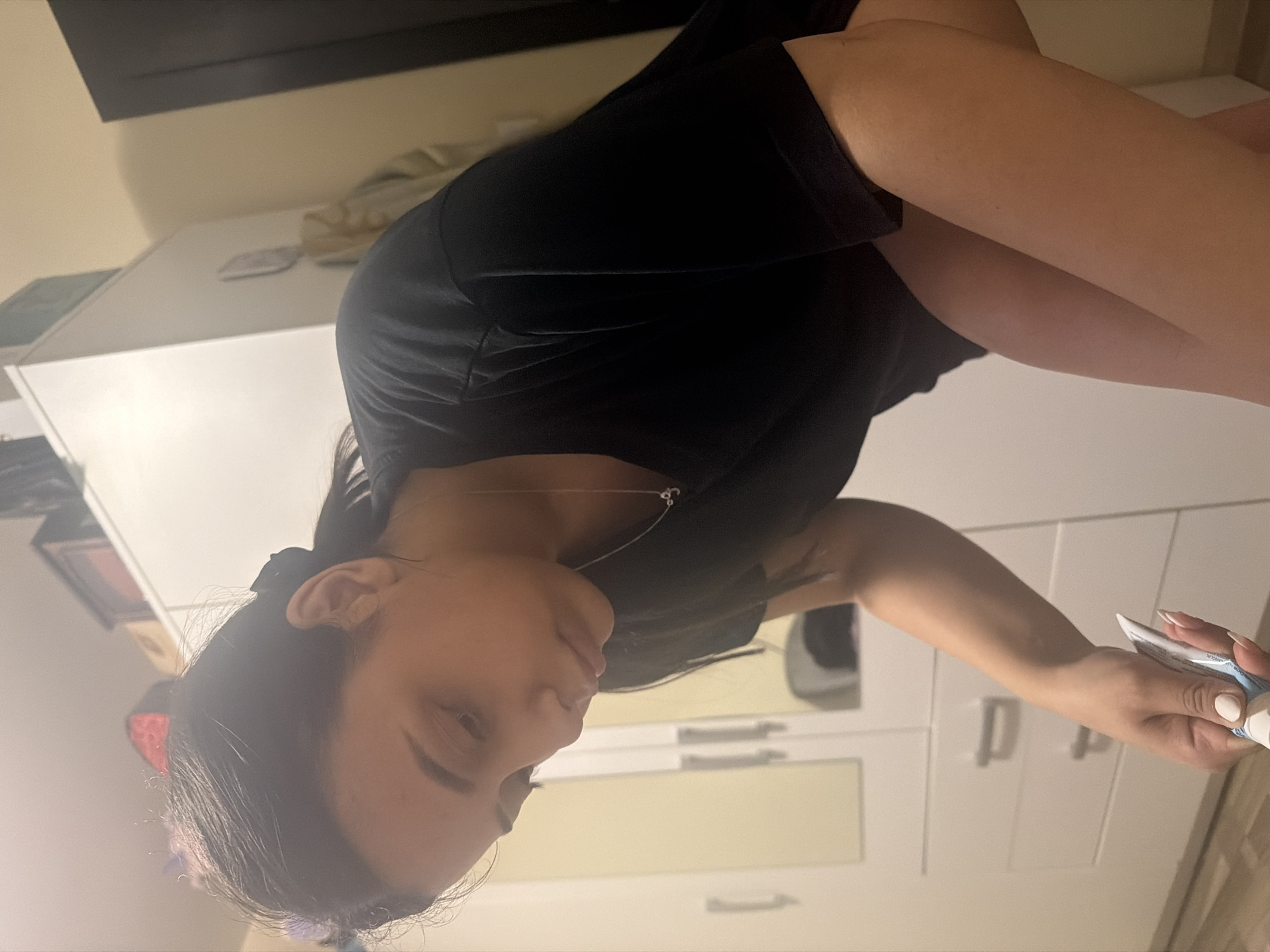 The image depicts a young woman in an indoor setting, likely in Shumen, Bulgaria. She is seen from a high-angle perspective, appearing to be lying down or leaning back, with her head turned slightly to her left and looking upward. She has dark hair, styled away from her face, and is wearing a simple black t-shirt with a thin silver necklace. Her expression is neutral and relaxed.
In her right hand, which has white-painted fingernails, she holds a white and blue tube, possibly containing toothpaste or cream. The tube features some indistinct text, with fragments resembling "AR" and "OA" visible. Her left arm is bent.
The background consists of light-colored walls and a modern white dresser or cabinet with multiple drawers or doors, indicated by visible handles. Above the dresser, a dark, flat object, possibly a TV or a dark shelf, spans the upper portion of the frame. The lighting is artificial, illuminating the scene without specific cues about the time of day, suggesting an indoor activity. The overall impression is one of a calm, personal moment.