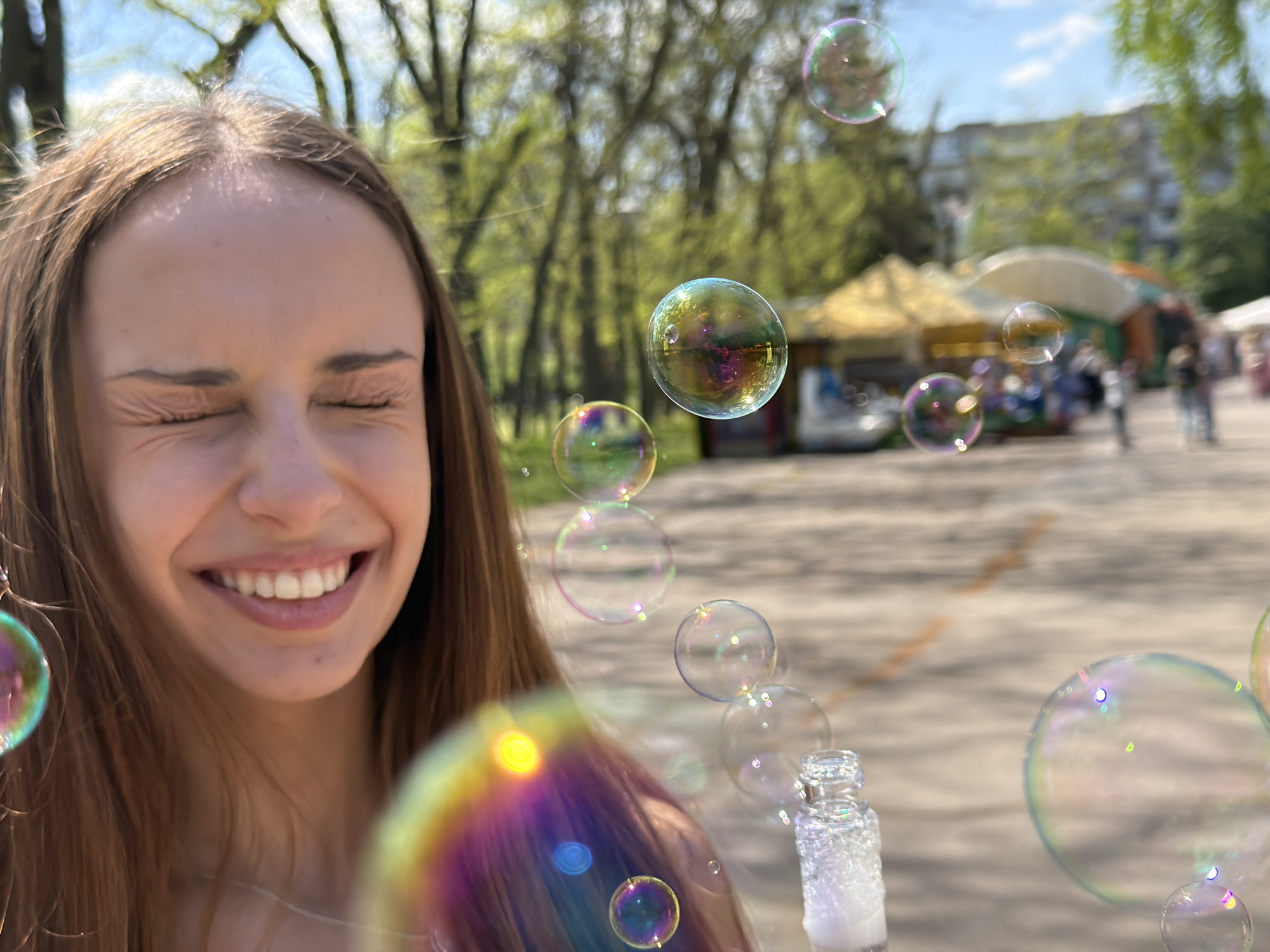 The image shows a young woman with long brown hair, eyes closed and smiling, surrounded by soap bubbles in a park. She is holding a bottle with bubble liquid. The background reveals a blurred view of a park or outdoor fair, with trees, tents, and people in the distance. The photo appears to be taken outdoors on a sunny day. The location is most likely in the city of Sofia, Bulgaria.
