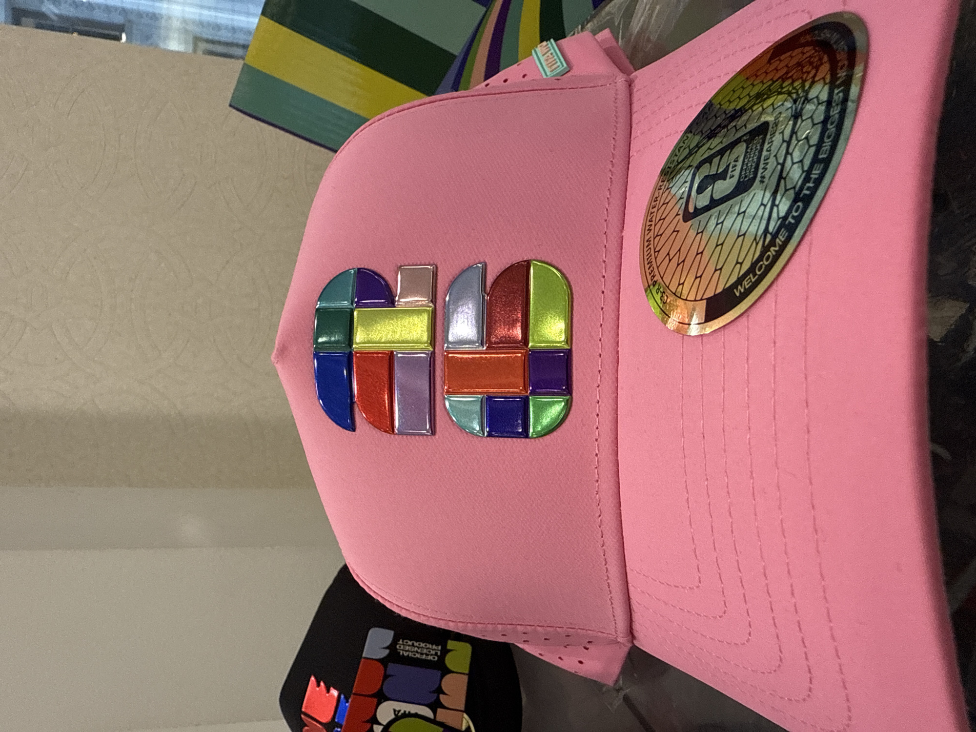The image shows a pink baseball cap with a colorful mosaic-style design on the front, resembling block letters. There's also a circular sticker on the side of the cap with FIFA branding and text that reads "WELCOME TO THE BIGGEST MOMENT". A portion of another baseball cap with a similar mosaic design is visible underneath, partially obscured. The background is nondescript, with hints of beige and a green-yellow striped object in the upper background. Based on the branding and design, it's likely that these caps are related to the 2026 FIFA World Cup, which will be hosted in part in Chicago, United States of America.