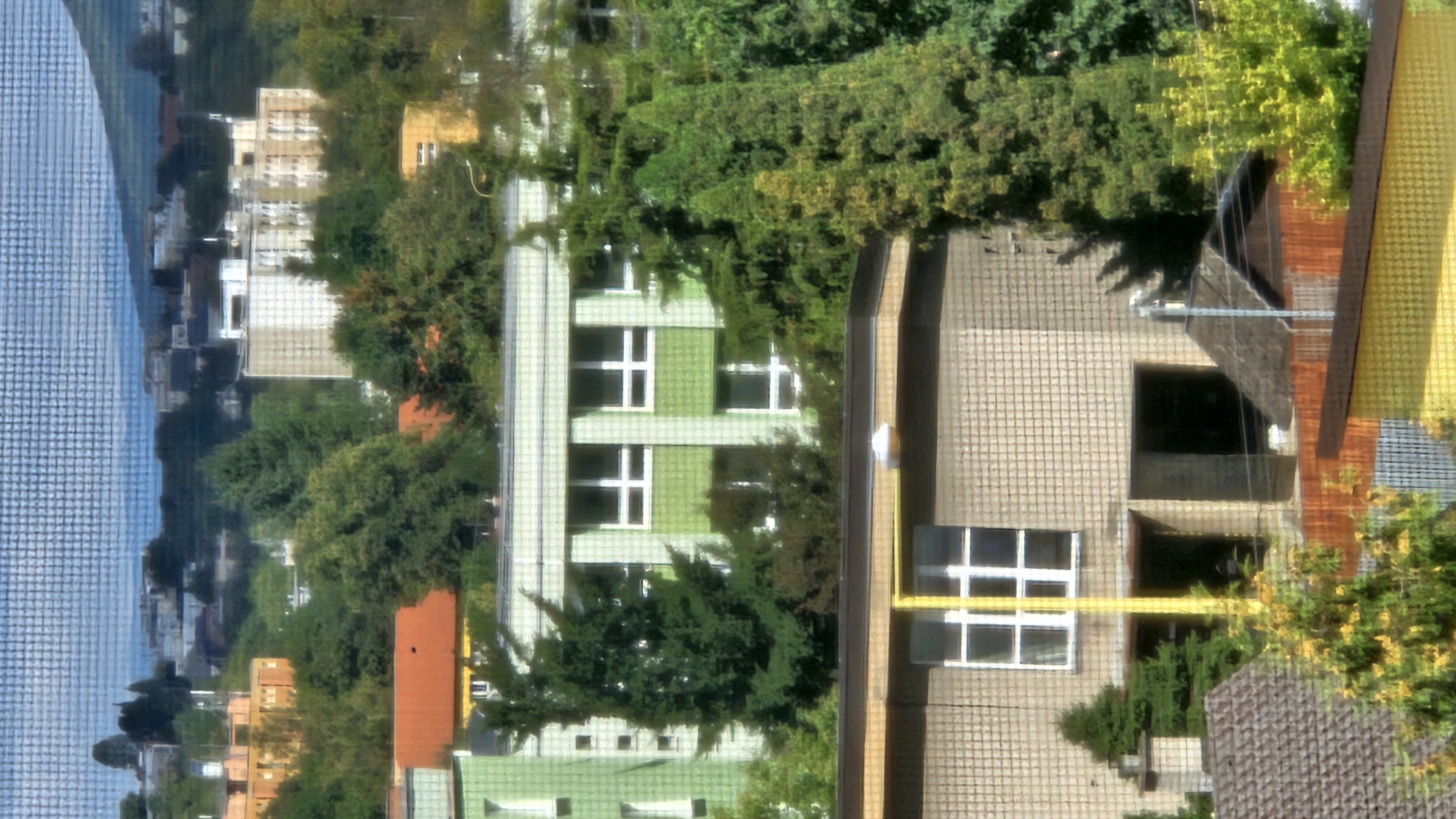The image captures a view of residential buildings and greenery in the city of Prešov, Slovakia. In the foreground, a building with a beige tiled facade features several windows, one of which is prominently divided into panes by a white grid. A vertical yellow pole extends upwards in front of this building. Partially visible behind this structure is another building with a green tiled exterior and white-framed windows. Further in the background, more buildings of varying heights and colors, along with lush trees and vegetation, are discernible. The scene is set against a backdrop of a hazy sky and distant rolling hills.