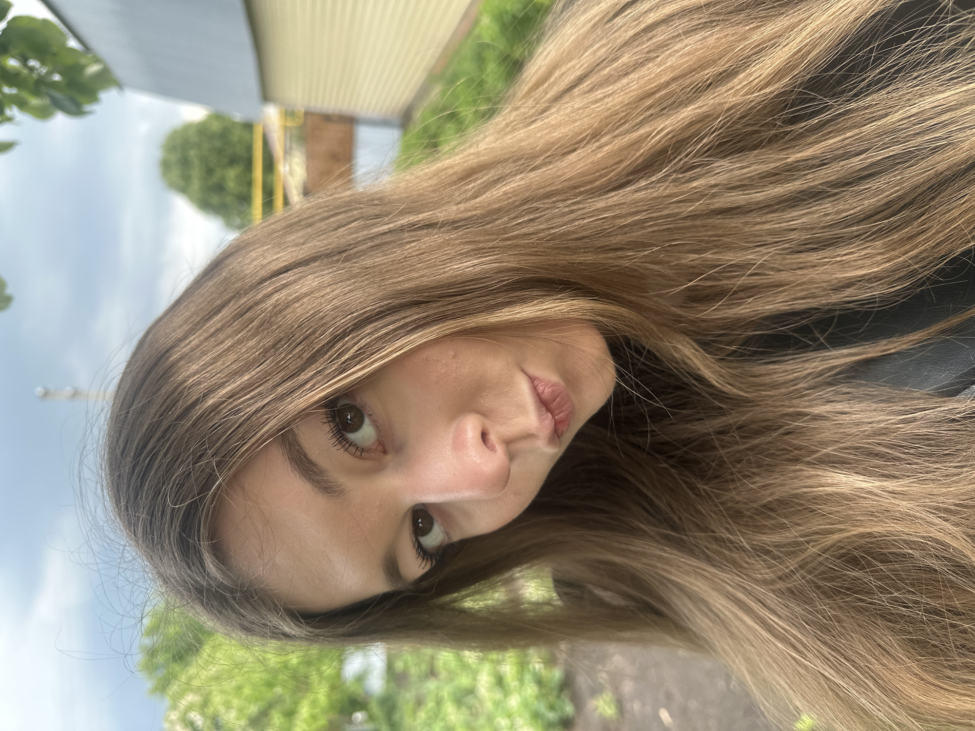 A young woman with light brown hair is looking at the camera in an outdoor setting. Green foliage and trees are visible in the background, along with a light-colored structure possibly made of metal. The image appears to be taken in Коноваловка, Russia. The lighting is soft, suggesting it might be an overcast day.