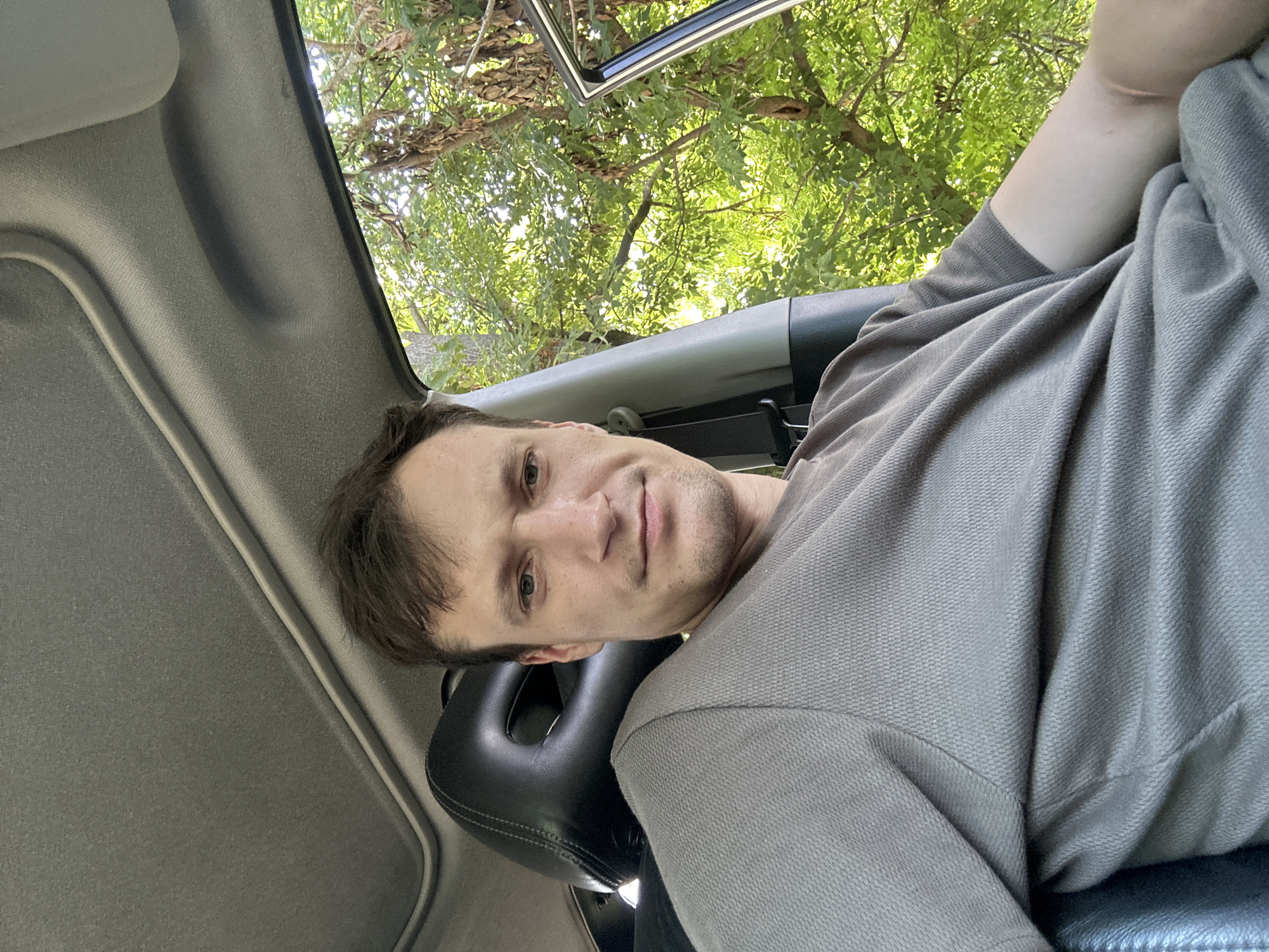 A man is sitting in the driver's seat of a car, with a sun roof open above him. He is wearing a grey t-shirt. The car is parked outdoors, with lush green trees and foliage visible through the open car door and windows. The background appears to be a natural setting, possibly a park or a rural area. The man is looking at the camera and appears to be relaxed. The lighting suggests it is daytime. The specific location in Makiv, Ukraine is not determinable from the image alone.