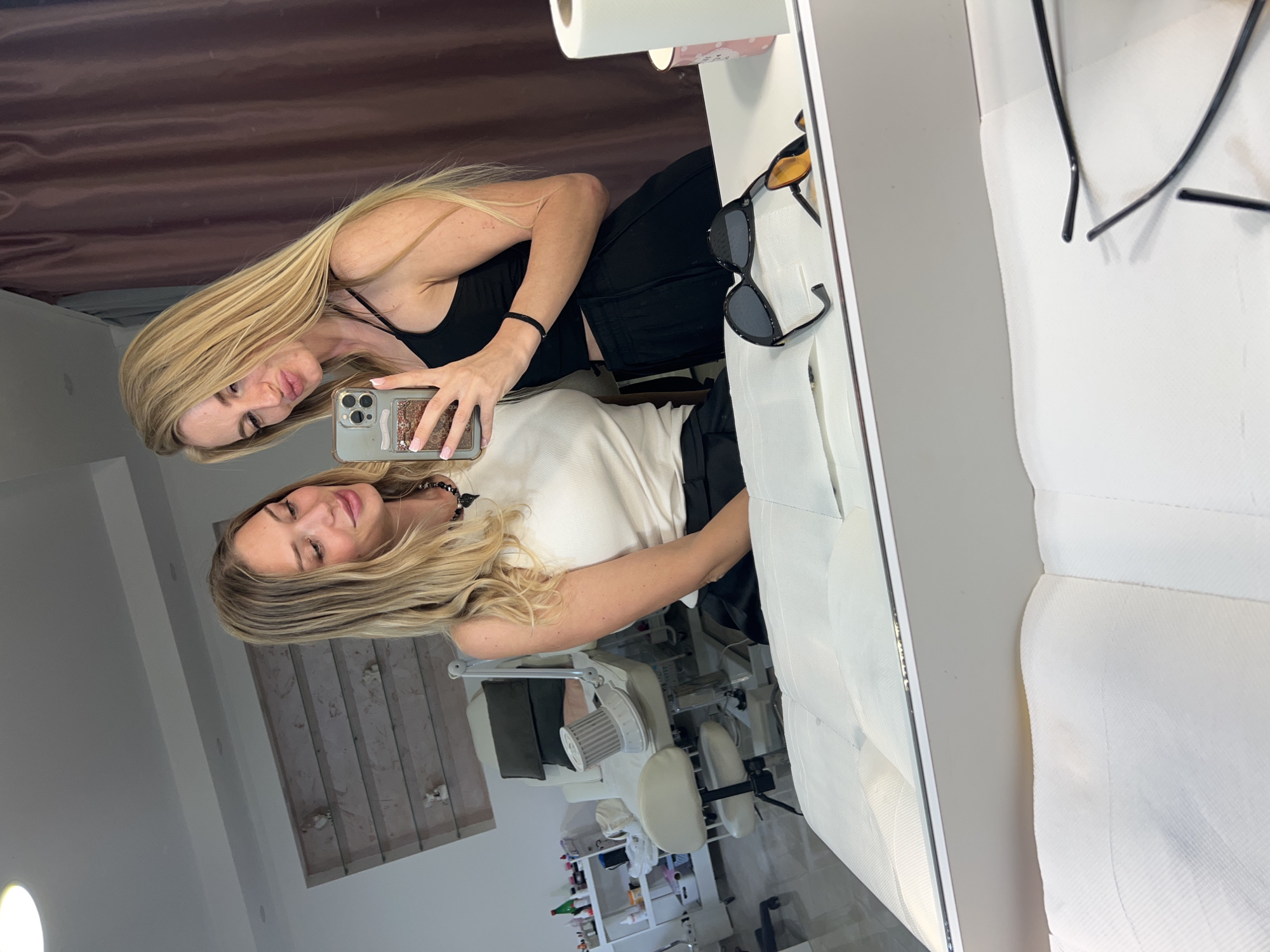 Two blonde women are reflected in a mirror. One woman, in a black tank top, holds a cell phone taking a photo. The other woman, in a white top, stands beside her. The mirror reflects a room with a salon chair and medical or salon supplies on shelves. The location may be a salon or clinic in Budva, Montenegro. Sunglasses are placed on a shelf next to the mirror. A roll of paper is on the shelf as well.