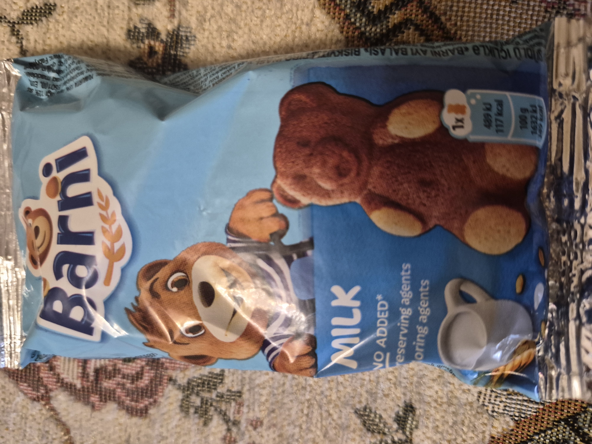 The image shows a close-up of a bag of "Barni" milk biscuits. The bag is predominantly light blue and features cartoon illustrations of teddy bears. The brand name "Barni" is prominently displayed in large, blue letters at the top of the bag. Below the brand name, a teddy bear character wearing a striped sailor shirt is depicted. Further down, the word "MILK" is written in large, white letters, followed by "NO ADDED*" and text that appears to list ingredients or nutritional information. A second, larger teddy bear-shaped biscuit is shown on the right side of the bag, alongside a nutritional information graphic indicating 1x biscuit, 489 kJ, 117 kcal, and 100g. The bag is resting on a patterned fabric surface, which is textured and appears to be a blanket or tablecloth. The lighting suggests an indoor setting, possibly a home environment, with a slight glare on the plastic packaging. The image does not contain any information that would place it in the city of Yerevan, Armenia.