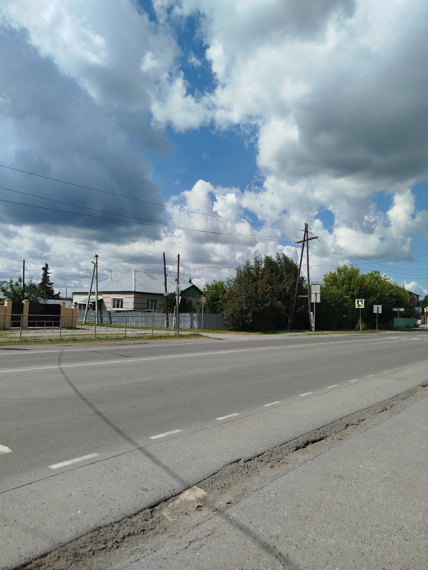 The image depicts a roadside view in Zavodoukovsk, Russia, under a dynamic sky filled with both bright blue patches and large, dark, scattered clouds. The foreground is dominated by a paved asphalt road with visible white lane markings. A portion of the road on the right shows signs of wear and damage along the edge.
Across the road, a low fence made of brick and metal separates the roadway from a residential area. Behind the fence, a single-story house with a light-colored metal roof is visible, constructed with white brick. Various trees and dense foliage line the property, with some appearing to be deciduous and others coniferous. Electrical poles with wires are present along the roadside, suggesting utility infrastructure.
Further down the road, on the right side of the frame, a pedestrian crossing sign is prominently displayed on a yellow square with a black border and a black pedestrian symbol inside. This indicates a designated crossing point. The overall scene suggests a suburban or semi-rural environment within Zavodoukovsk, characterized by residential buildings, basic infrastructure, and a natural landscape. There are no people visible in the image.