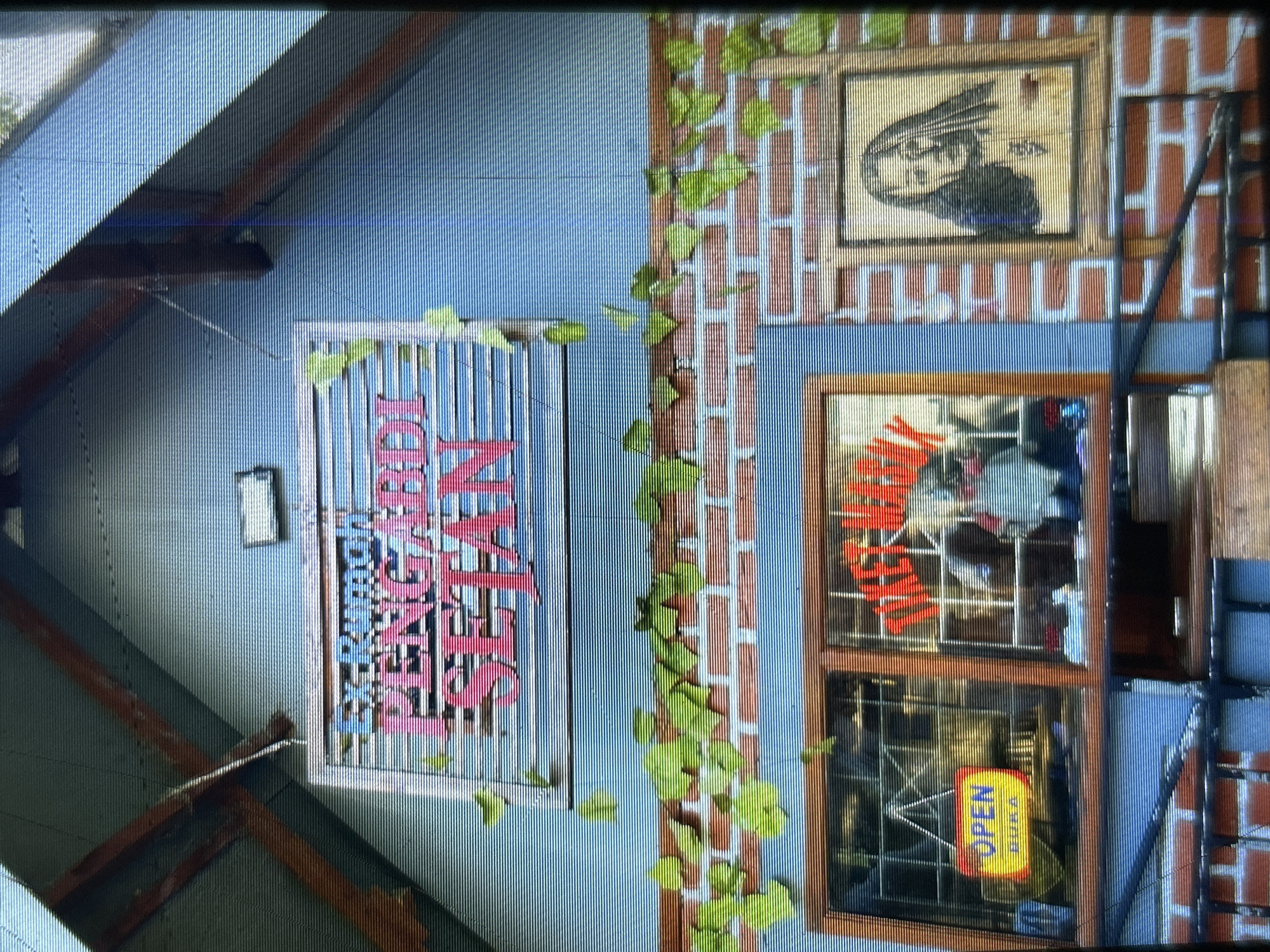 The image captures the facade of a building in Depok, Indonesia, with a distinctly vintage and somewhat eerie aesthetic. Above a brick-patterned wall, a sign with pink lettering reads "Rumah Pengabdi Setan," which translates to "House of Satan Worshippers." This sign is positioned on a blue wall with wooden beams supporting a slanted roof. Green foliage drapes over the brickwork, adding a touch of nature to the scene.
Below the sign, there are two windows. The left window displays an "OPEN" sign with yellow and blue text, suggesting it's an entrance to a shop or establishment. The right window appears to be a display area, with red neon lettering that is partially obscured, but the word "TIKET" (ticket) is visible. To the right of the windows, a framed portrait of a woman with long, dark hair is mounted on the brick wall. The overall impression is that of a themed attraction or an establishment with a gothic or horror-inspired motif. The image exhibits some visual distortion, likely due to the camera's lens or recording medium, characterized by horizontal lines and a slightly grainy texture.