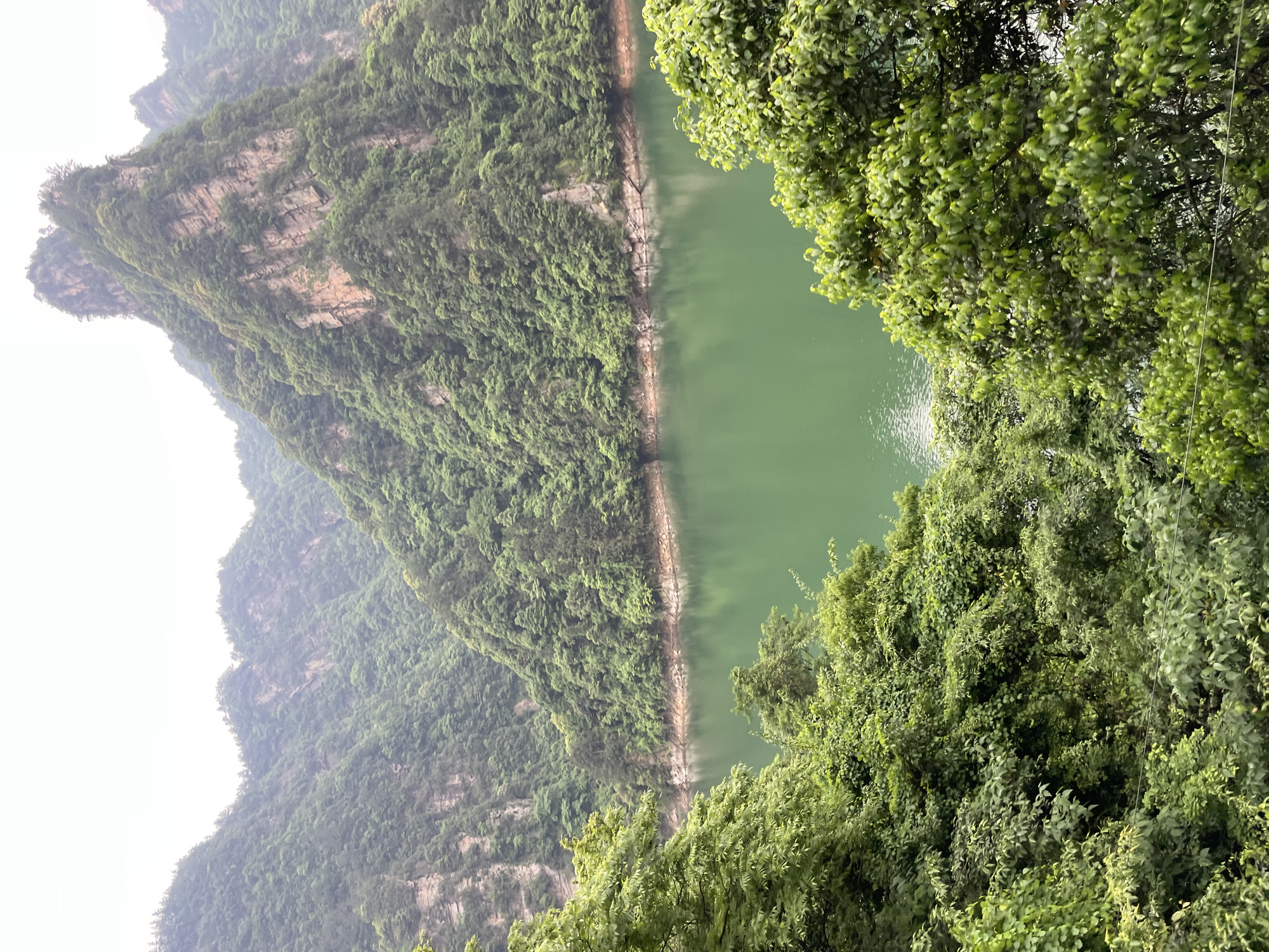 This image shows a scenic view of the Zhangjiajie National Forest Park in Wulingyuan, China. A body of water, likely a river or lake, is positioned between steep, verdant mountains. The mountain on the left has exposed rock formations. Lush green foliage and trees surround the water and dominate the foreground of the image. The weather appears misty or overcast, contributing to the landscape's ethereal quality.