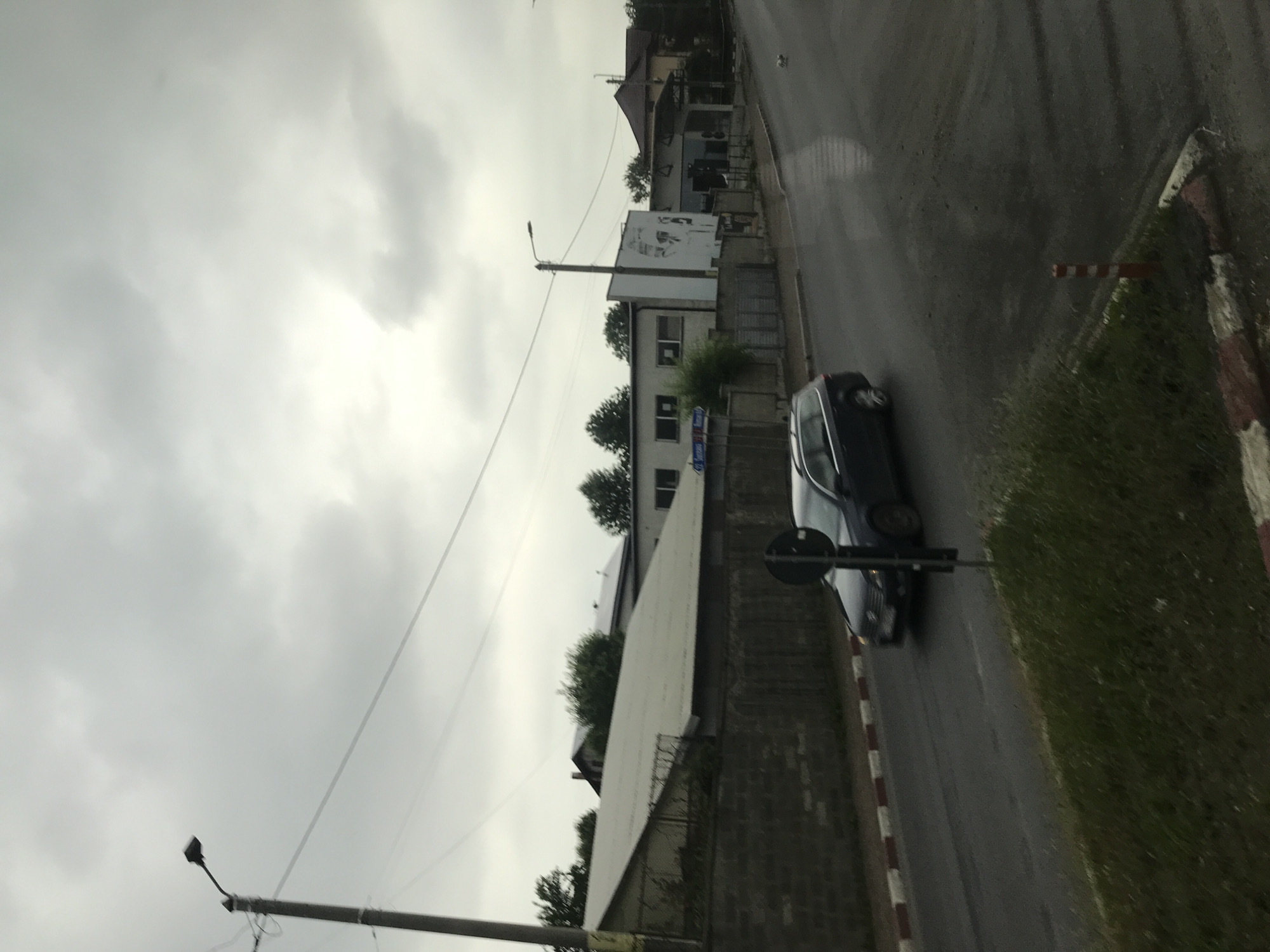 This image depicts a street scene in Fălticeni, Romania, identified by a blue street sign indicating "Strada Suceava" and "Strada Mioriței" on a building's fence. The scene is captured under an overcast, grey sky.
A dark-colored station wagon is visible in motion, traveling from left to right on a wet asphalt road, suggesting recent rainfall. The road is bordered by red and white striped curb markings.
On the left, a prominent utility pole with a streetlamp stands, from which power lines extend across the upper-mid section of the frame. Behind a low stone wall on the left, a large building with a light-colored, gently sloping roof is visible. To its right, a two-story, light-colored building features multiple windows, with the aforementioned street sign attached to its perimeter fence. Adjacent to this building is a large white billboard or wall structure displaying a stylized black-and-white graphic of a face. Further down the street on the right, another utility pole with a streetlamp is present. Residential buildings with reddish-brown tiled roofs are discernible in the background.
In the foreground, patches of green grass are visible. A black circular sign on a pole is positioned in the lower-center left. On a grassy median in the lower right, a small red and white striped bollard is present. No people are visible in the scene.