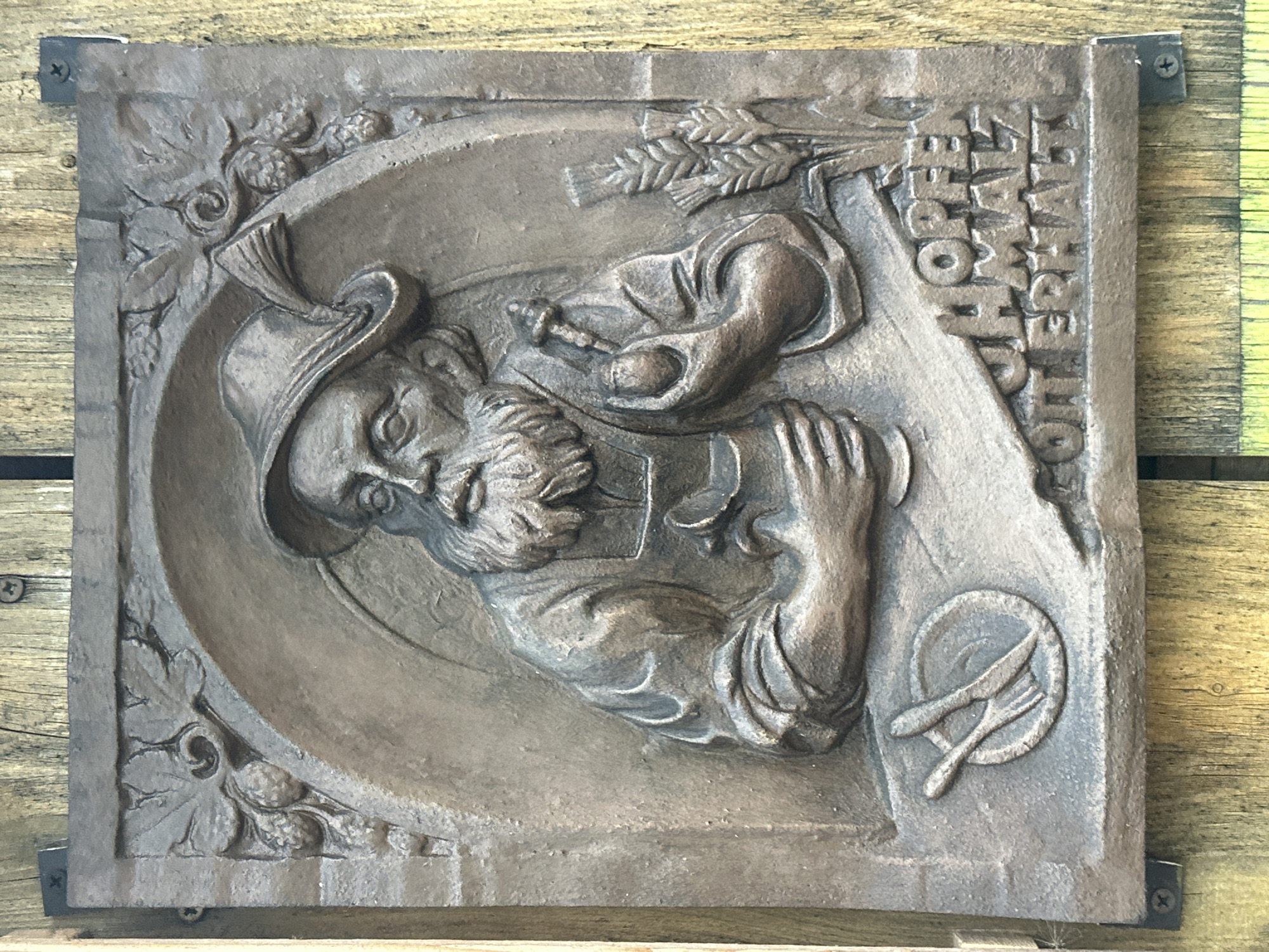 The image is a close-up, eye-level, outdoor photograph of a weathered bronze relief plaque mounted on a wooden fence. The plaque depicts a man with a beard and a feathered hat, holding a pipe and a mug, with a fork and knife on a plate to his left, and wheat stalks to his right. The text "HOPFEN MALZ GOTT ERHALT" is inscribed on the lower right of the plaque. The plaque is framed by grapevines at the top. The wooden fence behind it has visible grain and is weathered. The lighting suggests it is daytime.