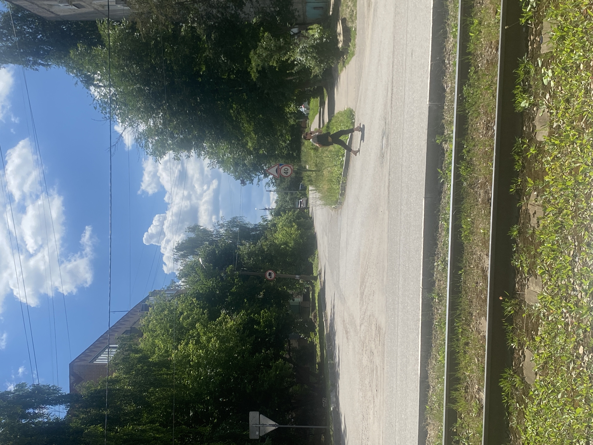 A person is skateboarding down a paved road in Kolomna, Russia. The scene is bathed in sunlight, suggesting it is daytime. The sky is blue with fluffy white clouds, indicating pleasant weather. Lush green trees line the sides of the road, with some buildings visible in the background.
The skateboarder is a young person wearing dark pants and a light-colored shirt. They are in motion, their body angled as if balancing on the board, with one leg extended behind them.
Several traffic signs are visible along the road. One sign shows a red circle with a diagonal line through it, indicating a prohibition, and a vehicle symbol inside. Another sign shows the number "40" within a red circle, likely indicating a speed limit. A triangular sign with a white background and a red border is also present.
The road appears to be a typical urban street, possibly a residential area, with a grassy verge and some overgrown vegetation. The overall impression is a casual, outdoor activity on a clear day.