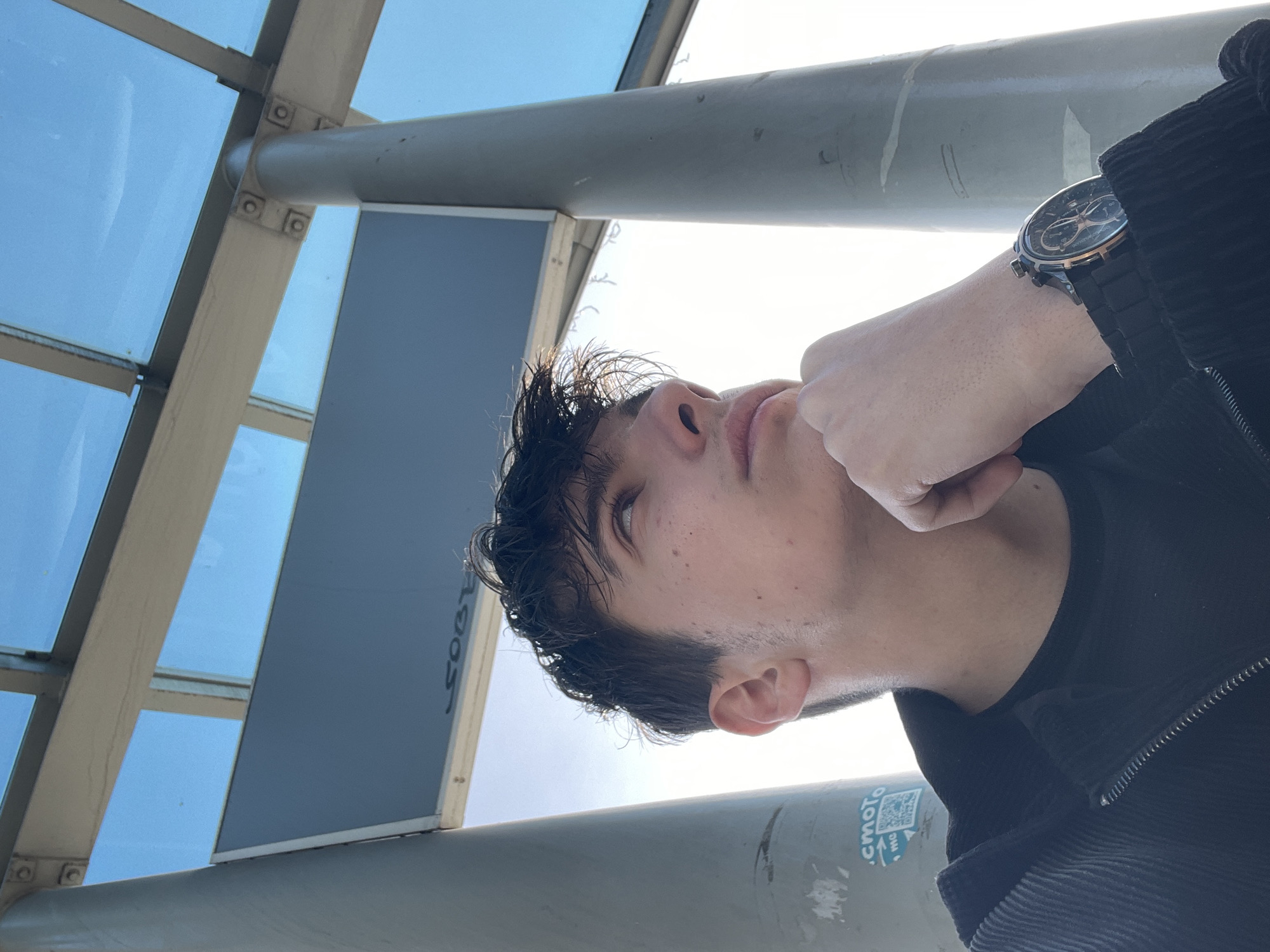 A low-angle shot depicts a person looking upwards, with their hand near their chin and a wristwatch visible on their wrist. The background consists of a metal structure with glass panels, suggesting an outdoor urban environment. A sign is partially visible behind the person's head. A sticker on the metal pole in the lower right corner of the image contains a QR code and some partially visible text, potentially "Burgas". Based on the sticker with the QR code on the metal pole, the location can be identified as being in Burgas, Bulgaria. The person is wearing a dark-colored jacket.