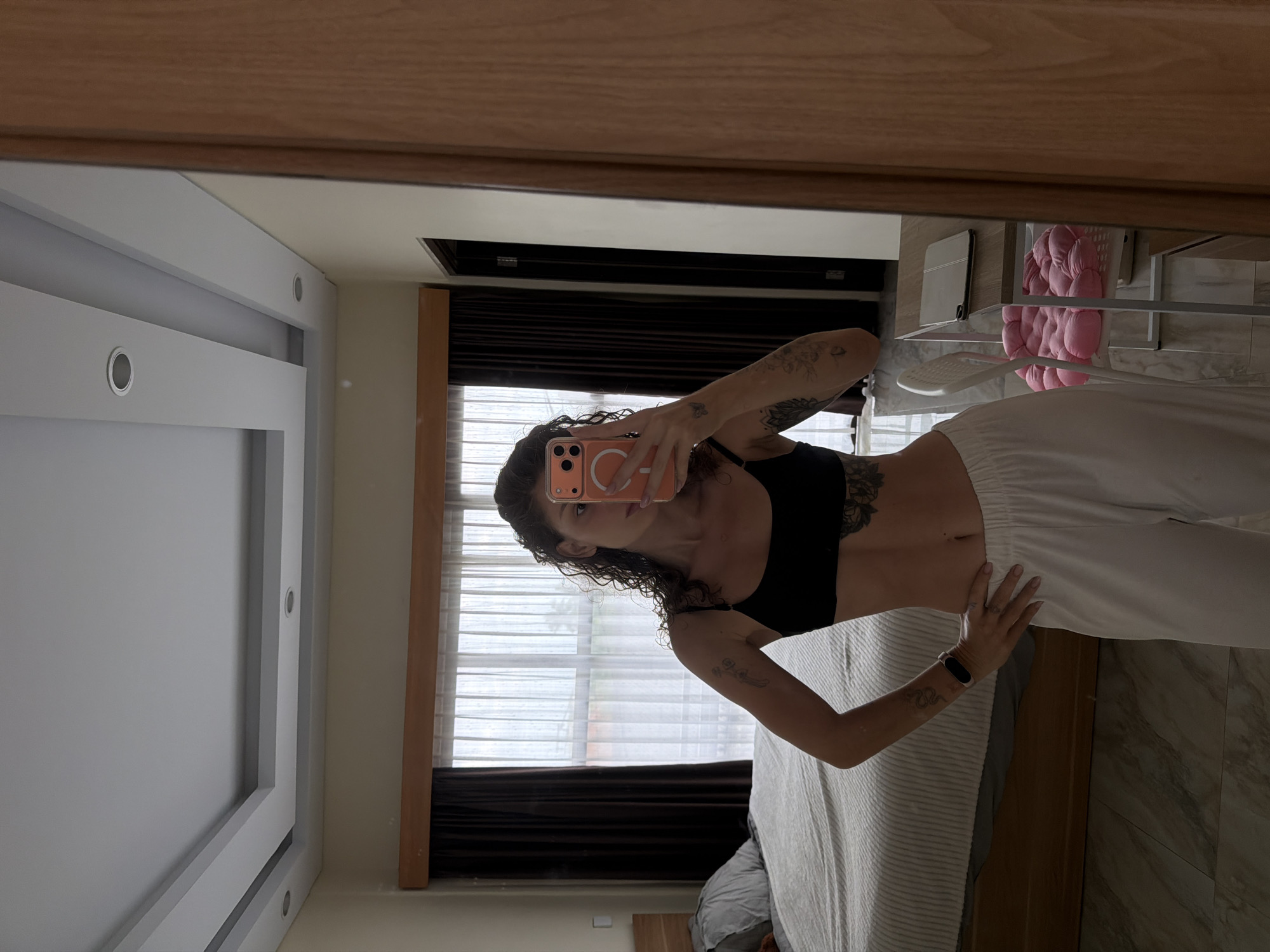 In a bedroom located in North Kuta, Indonesia, a woman is taking a mirror selfie. She is standing in front of a large mirror, holding her phone up to capture her reflection. The woman is wearing a black sports bra and white pants. She has curly brown hair and several tattoos on her arms and torso. Her left hand is on her hip, and her right arm is extended, holding the phone.
The bedroom is furnished with a bed, a wooden headboard, and a nightstand. The bed is covered with a striped comforter and a grey pillow. Behind the bed, there is a large window with sheer white curtains and dark brown drapes. The room has a modern ceiling with recessed lighting. To the right of the woman, there is a desk with a laptop and a chair with a pink cushion. The overall impression is of a contemporary and well-lit living space.