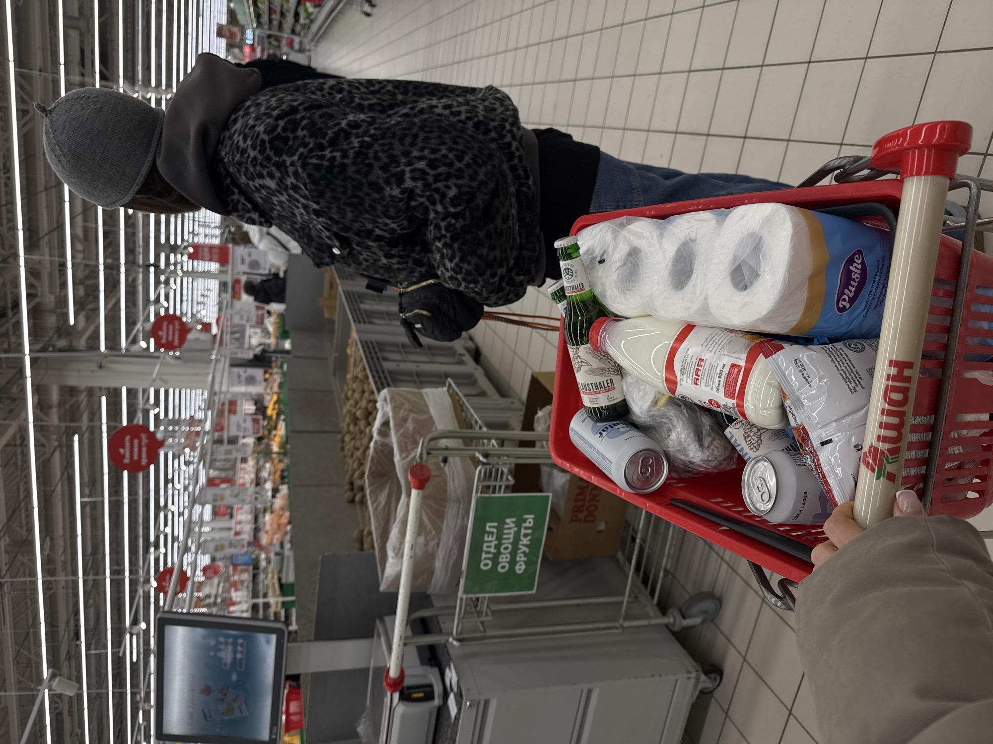 The image shows a shopping cart in a supermarket, likely in Moscow, Russia, based on Cyrillic text on signs and product labels. The cart is filled with items including toilet paper (a "Plushe" brand), bottles of milk, beer (possibly "Lausethaler" brand), and canned drinks. A person wearing a gray hat and patterned jacket is pushing the cart. The supermarket aisles are visible in the background with shelves stocked with various products. A green sign reads "ОТДЕЛ ОВОЩИ ФРУКТЫ", meaning "Vegetable Fruit Department." A digital information screen is visible near the bottom of the frame. The shopper appears to be in the vegetable section of the store.