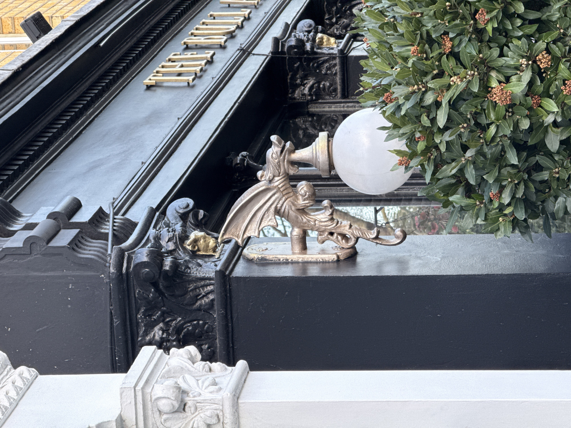 The image shows a close-up of architectural details on a building facade, likely in London, United Kingdom. The main subjects are ornate decorations, including a dragon-shaped light fixture mounted on a black surface. To the right, there is a round streetlight partially obscured by a leafy bush. The architectural style is intricate, featuring decorative moldings, carved details, and a white balustrade in the foreground. Gold lettering is visible on the building's upper section. There is no specific action or interaction depicted in the scene. The environment appears to be an urban setting, based on the architectural features.