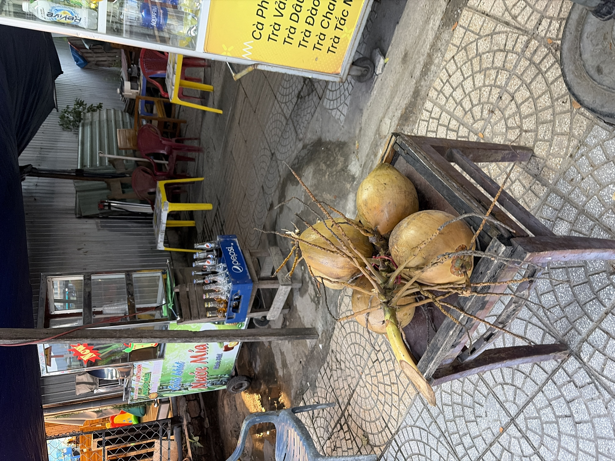 The image depicts an outdoor scene in the city of Đà Nẵng, Vietnam, likely in front of a small food or drink stand. The foreground is dominated by a cluster of coconuts resting on a weathered, dark wooden stool or crate. The coconuts are a light brown color and appear to be fresh, with visible brown fibrous husks and stems.
Behind the coconuts, a blue plastic crate filled with glass bottles of beverages, possibly beer or soft drinks, is visible. Some of the bottles have yellow liquid inside, and a Pepsi logo is clearly discernible on the crate. Further back, a mobile cart or stand is set up, featuring a metal structure with glass panels, likely for displaying products or equipment. The cart has signage with Vietnamese text, including "NƯỚC MÍA" (sugarcane juice).
The environment suggests a street-side setting. Paved ground with a circular tile pattern is visible. Several red plastic chairs and yellow plastic tables are arranged in the background, indicative of an outdoor seating area for customers. The overall lighting suggests it is either early morning or late afternoon. A dark blue tarp or awning covers part of the scene. The background wall appears to be corrugated metal. No people are visible in the frame.
