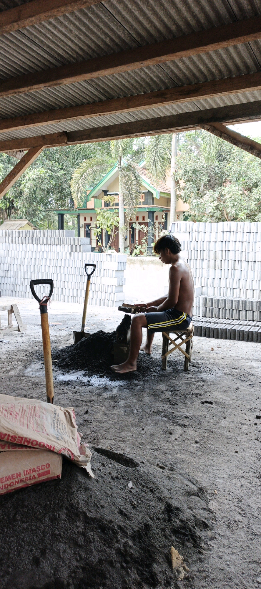 In Kasiyan, Indonesia, an unclothed male is seen from the side, sitting on a small, wooden stool. He is working with a pile of dark, granular material, likely cement and sand, which is spread on the ground. In front of him, a wooden block sits on the material, and he appears to be using a tool, possibly a small trowel or brick mold, to shape or compress the mixture. Two shovels are positioned in the pile, one with a black handle and a wooden shaft with a red band, and another with a wooden handle and a black head. To the left of the shovels, a large, light-colored bag, partially open, is visible. The bag has red lettering that appears to say "IMASCO INDONESIA". Behind the male, stacks of gray, rectangular bricks are neatly arranged. The scene is set outdoors under a corrugated metal roof supported by wooden beams. In the background, a two-story house with an orange roof and green trim is visible, surrounded by trees, including palm trees. The ground is dirt and concrete.
