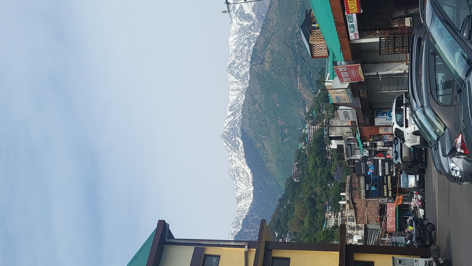 The image depicts a view of a street in Dharamshala, India, with snow-capped mountains visible in the background. The foreground shows buildings, commercial establishments with signboards in Hindi and English, parked cars and motorbikes. The sky is light blue with minimal cloud cover. The mountains are green on the lower slopes, transitioning to snow-covered peaks.