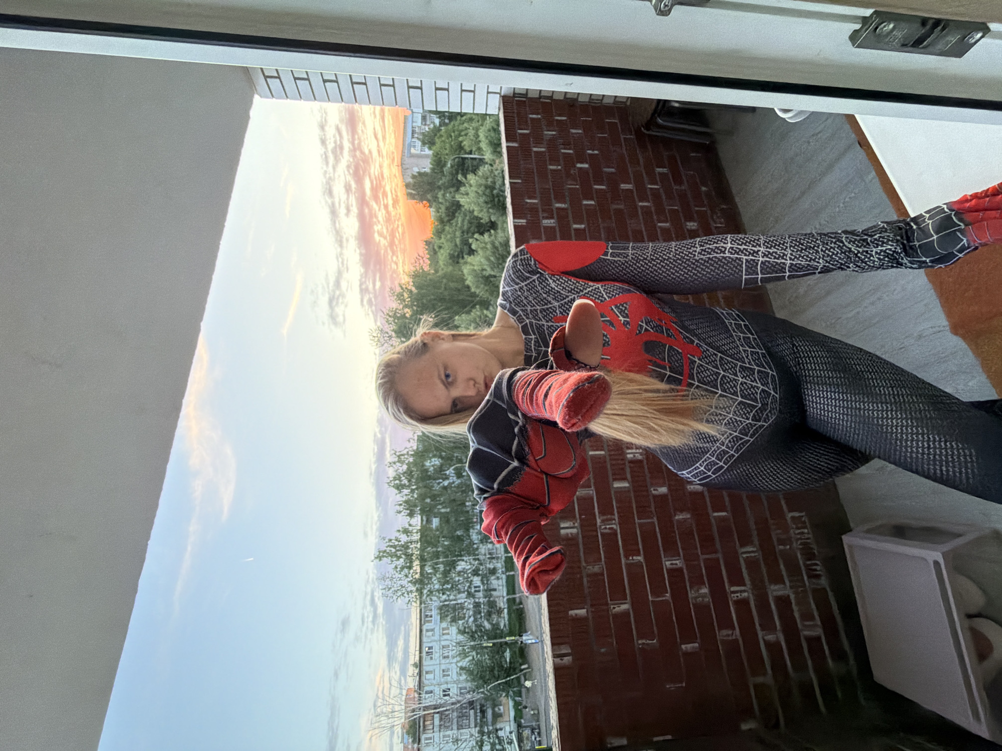 A young woman in a Spider-Man costume stands on a brick balcony, looking at the camera and holding up her hands. The sunset colors the sky. A city landscape with buildings and trees is visible in the background, which corresponds with the general architecture and urban layout of Izhevsk, Russia.