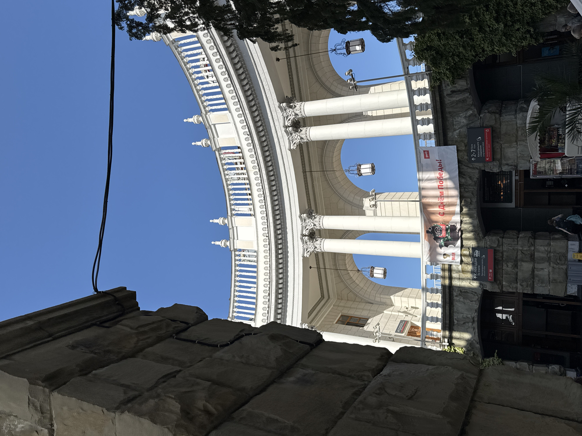 The image, taken from a low angle, features a portion of the Sochi railway station in Sochi, Russia. The building is ornate with white colonnades, balustrades, and decorative lamps. A banner with text in Russian hangs between the columns. In the foreground, there is a stone structure. Signage near a doorway indicates train ticketing information, and an "RZhD" logo is seen. The sky is blue and clear.