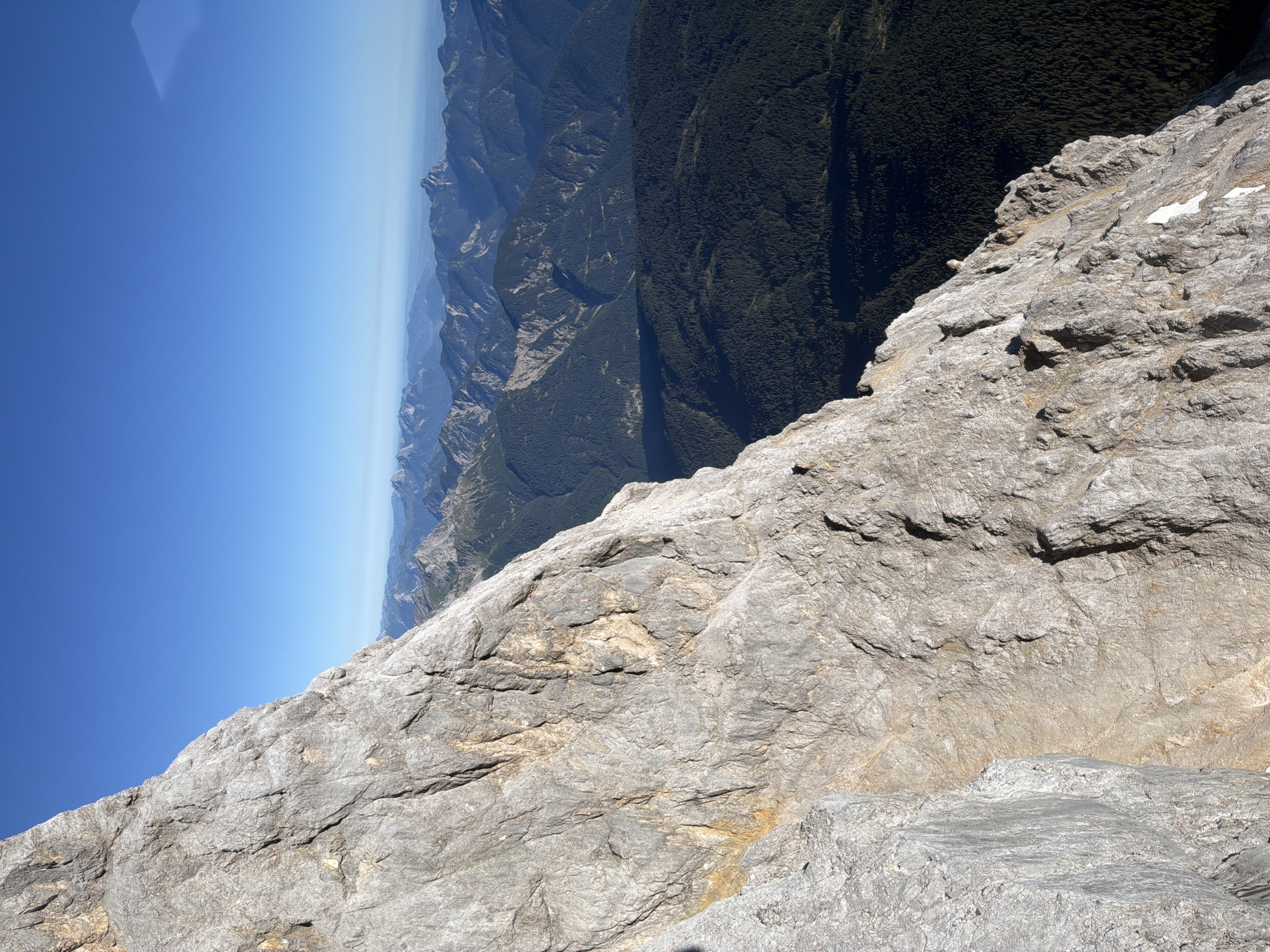 The image shows a high-altitude mountain landscape with a clear blue sky. In the foreground, a large, rugged, light-gray rock face dominates the left side of the frame, showing intricate textures and cracks. The rock face is angled, suggesting a steep slope or cliff edge.
Beyond the rock face, a vast expanse of mountain ranges stretches into the distance. The mid-ground is characterized by dark green, densely forested mountains, casting shadows. Further back, multiple layers of progressively lighter-hued mountain peaks are visible, fading into a hazy horizon. The overall scene depicts a remote, natural environment, likely within the Bavarian Alps, given the reference to Grainau, Germany, which is known for its proximity to the Zugspitze, Germany's highest peak. There are no people or specific man-made objects visible in the scene. The lighting suggests it is daytime, likely morning or afternoon, with the sun illuminating the rock face and the distant landscape.