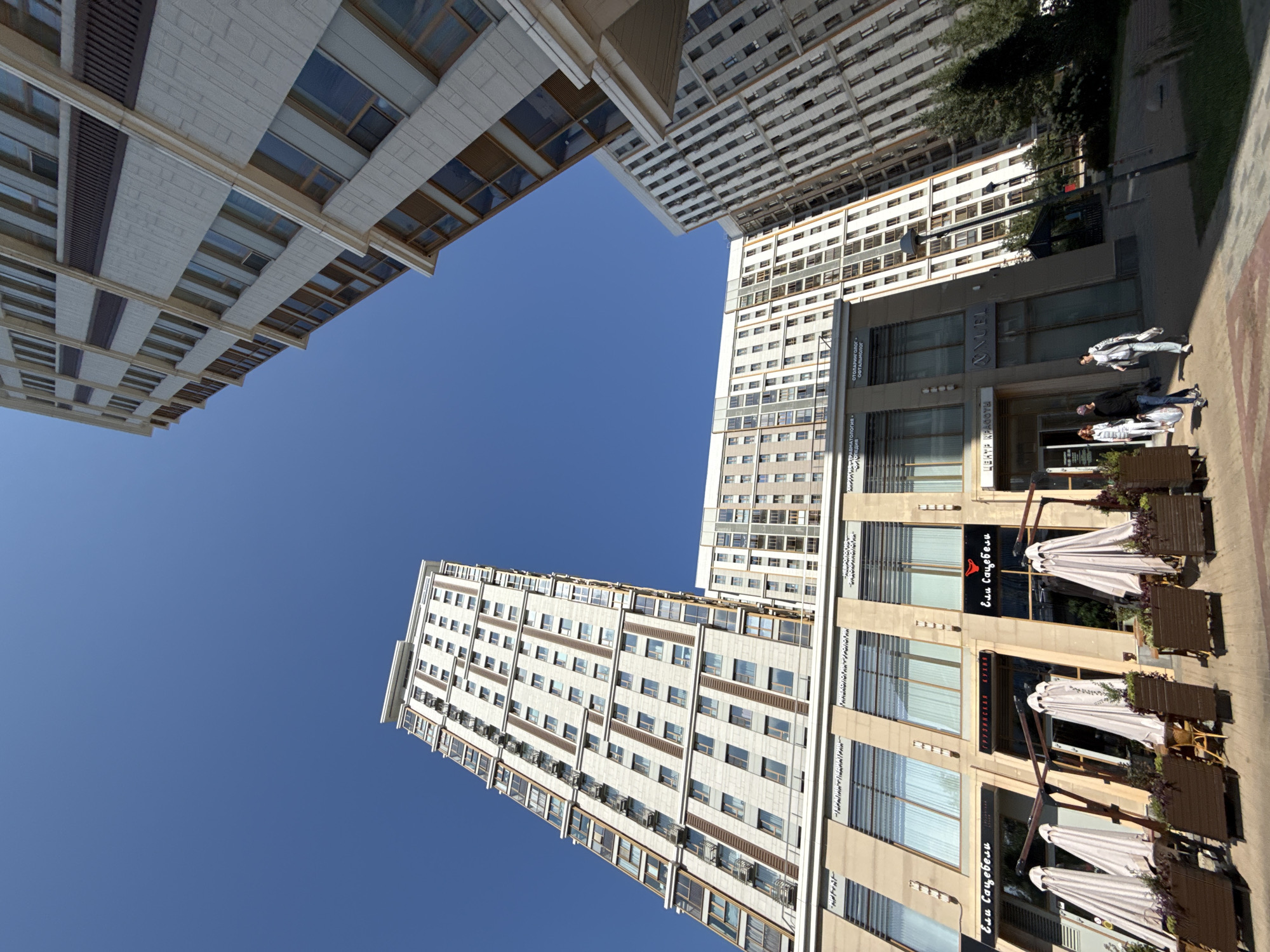 The image shows a modern residential complex with high-rise buildings and commercial spaces on the ground floor. The architecture is contemporary, with many windows and light-colored facades. The sky is clear blue, indicating a sunny day.
In the foreground, there are several outdoor seating areas with tables and umbrellas, suggesting a cafe or restaurant. Some planters with vegetation are also visible. Three people are walking on the sidewalk in front of the buildings. One person, wearing a black long-sleeved shirt and jeans, is carrying shopping bags. Another person, wearing a light-colored jacket and pants, is also carrying bags. A third person, wearing a white top and dark pants, is walking slightly behind them.
The signs visible on the buildings are in Russian. One sign reads "ЦЕНТР КРАСОТЫ" (Center of Beauty), and another appears to be for a Georgian restaurant, "Ели Сацебели" (Yeli Satsabeli).
The image captures a scene in a developed urban area, likely a residential district with amenities for residents and visitors. The perspective of the photograph emphasizes the height of the buildings and the vastness of the sky.
Based on the presence of the restaurant "Ели Сацебели" and the architectural style, this location is likely in Moscow, Russia. The specific complex appears to be "Art residence" (Арт Резиденс) or a similar modern residential development in the city.