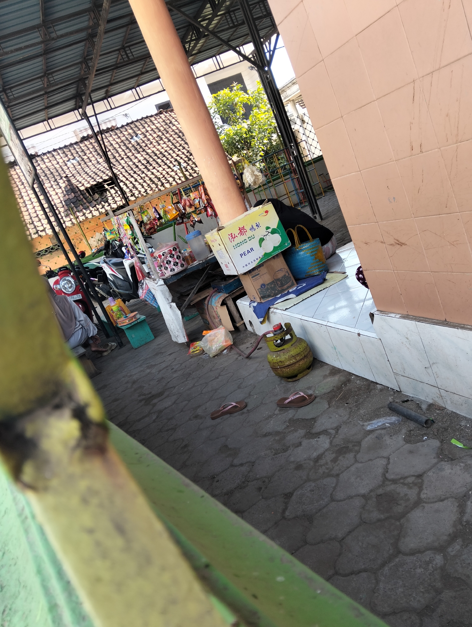 The image depicts an outdoor scene, possibly a market stall, in Muncar, Indonesia. A red motorcycle is parked to the left, with a person sitting on a bench nearby. In the foreground, a yellow propane tank rests on the paved ground next to a pair of brown flip-flops. Behind the tank, there is a tiled ledge with various items, including boxes, bags, and a plastic water bottle. Further back, a roof structure with tiled roofing is visible, supported by metal beams and a large, light-brown cylindrical pillar. The ground is paved with interlocking stones. The overall environment suggests a somewhat cluttered, utilitarian space often found in bustling market areas.