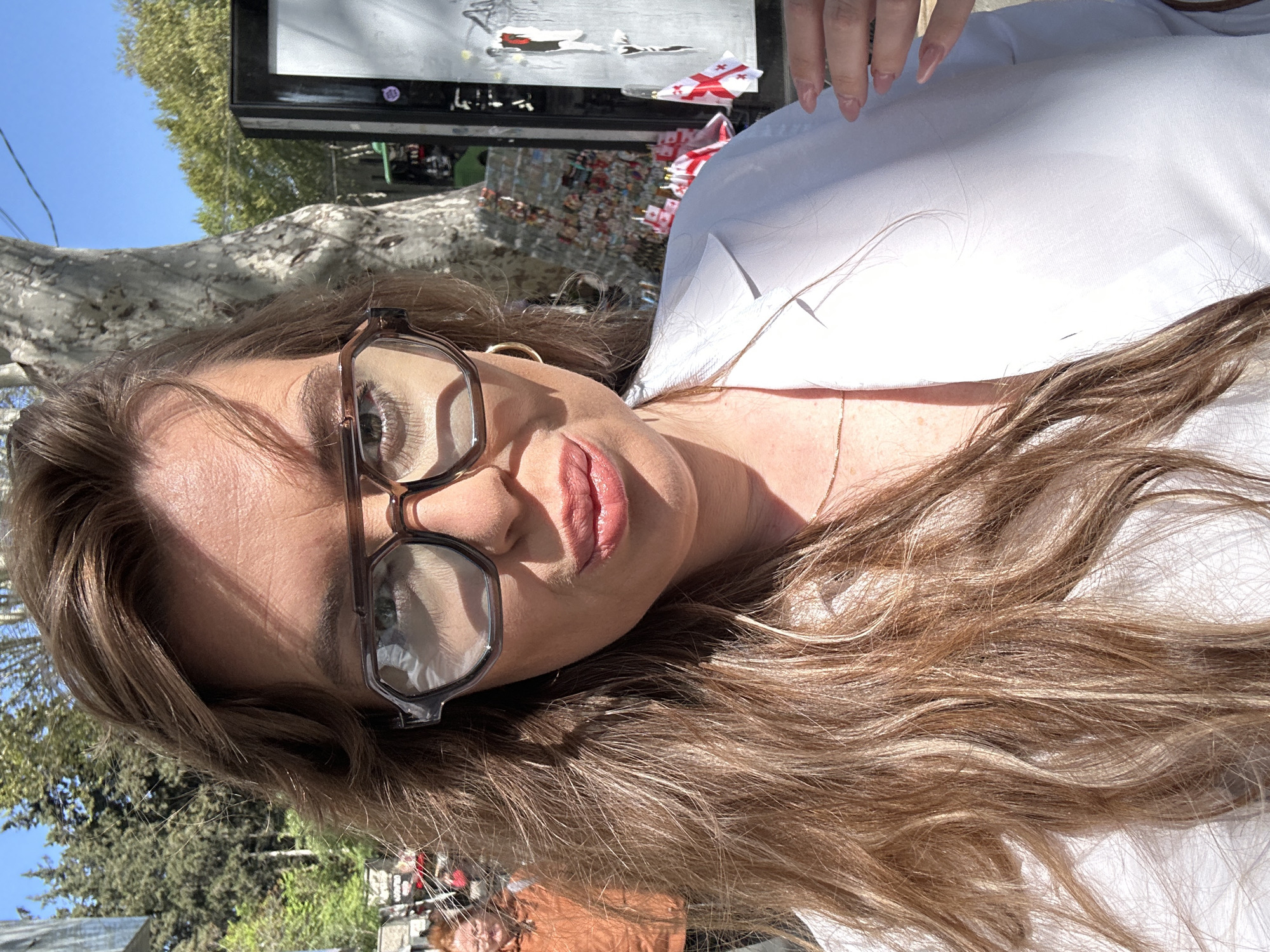 A close-up, portrait-style photograph, rotated counter-clockwise, features a woman looking directly at the camera. She has long, wavy light brown hair, which flows across her shoulder and chest, and is wearing a white collared shirt. She wears dark-framed glasses, and her lips are slightly parted with light pink lipstick. A thin gold chain is visible around her neck, and the fingers of her hand, with light pink manicured nails, are visible in the upper right.
The background suggests an outdoor setting under a clear blue sky with trees. Reflections in the upper part of the image, potentially in a window or display, reveal several small items. These include multiple small flags that prominently feature a white background with a red cross, identifying them as Georgian flags. Also visible are small models or decals of airplanes, one in white and another with red and white markings. The bright sunlight indicates it is daytime, casting clear shadows and highlights. The notable presence of Georgian flags strongly cues the location to be Tbilisi, Georgia. No clear, readable text is present.